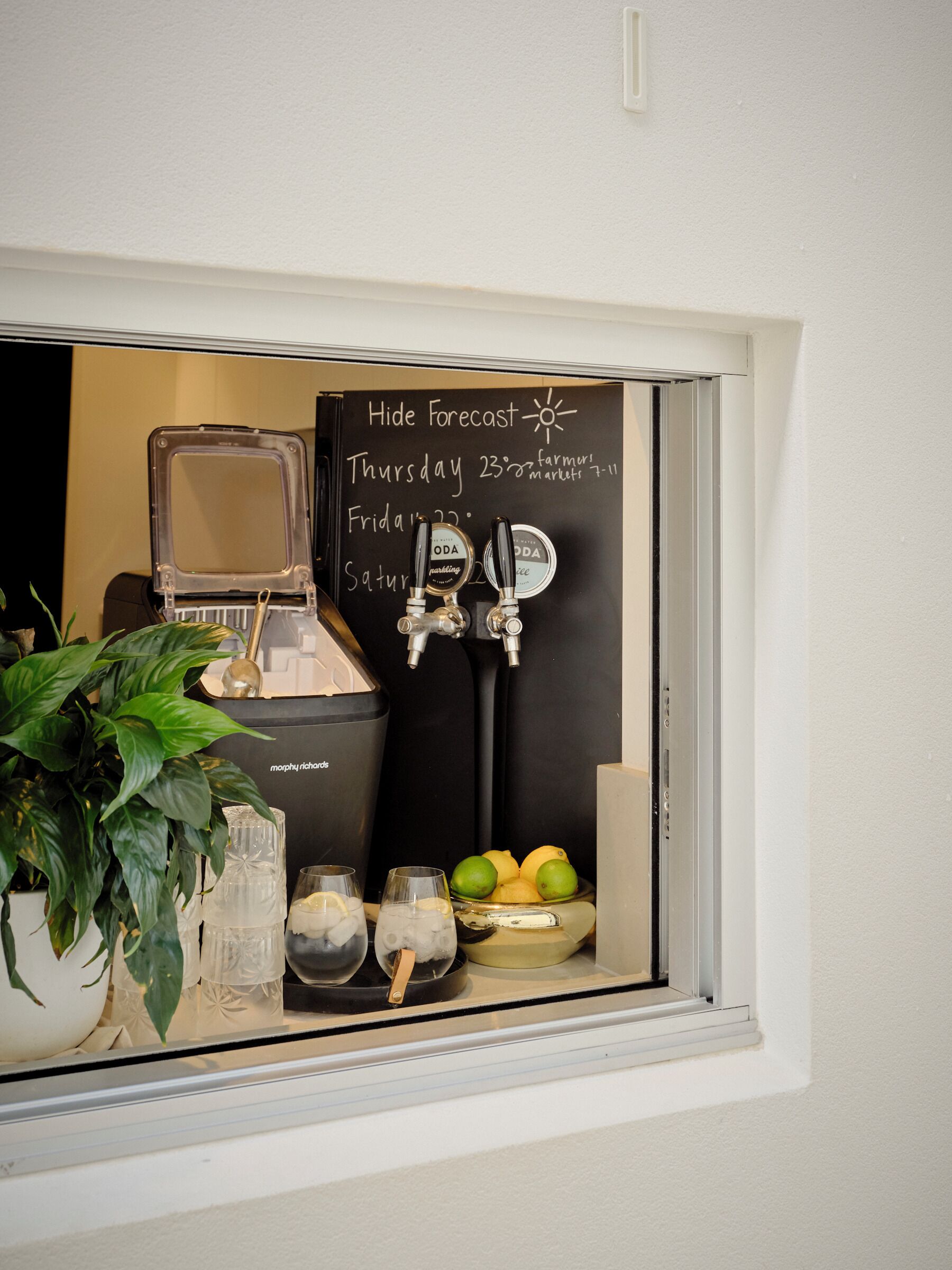 Una ventana con dispensador de agua, una pizarra con pronóstico del tiempo y un cuenco de fruta.