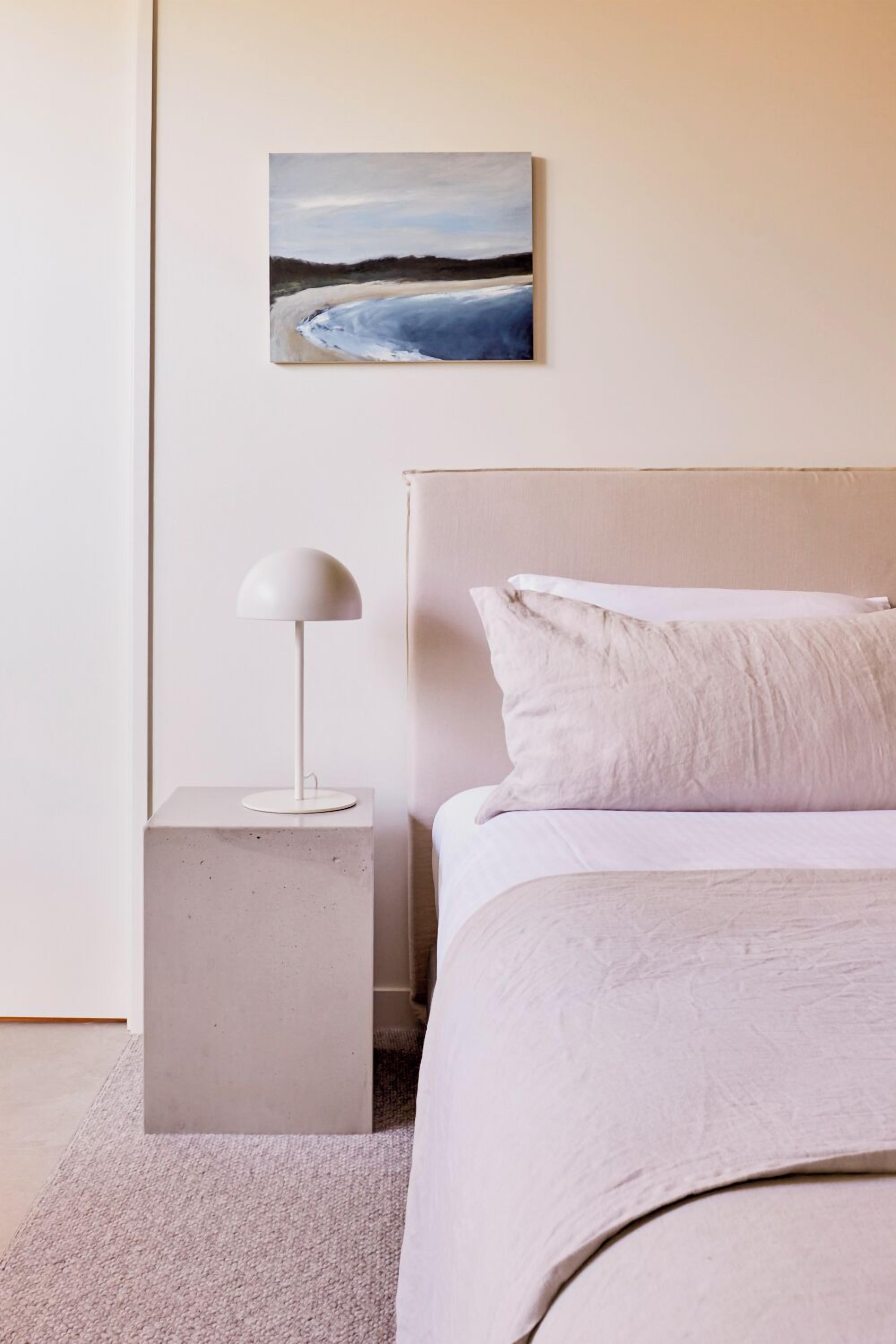 A minimalist bedroom with a bed, a nightstand, a lamp, and a painting on the wall.