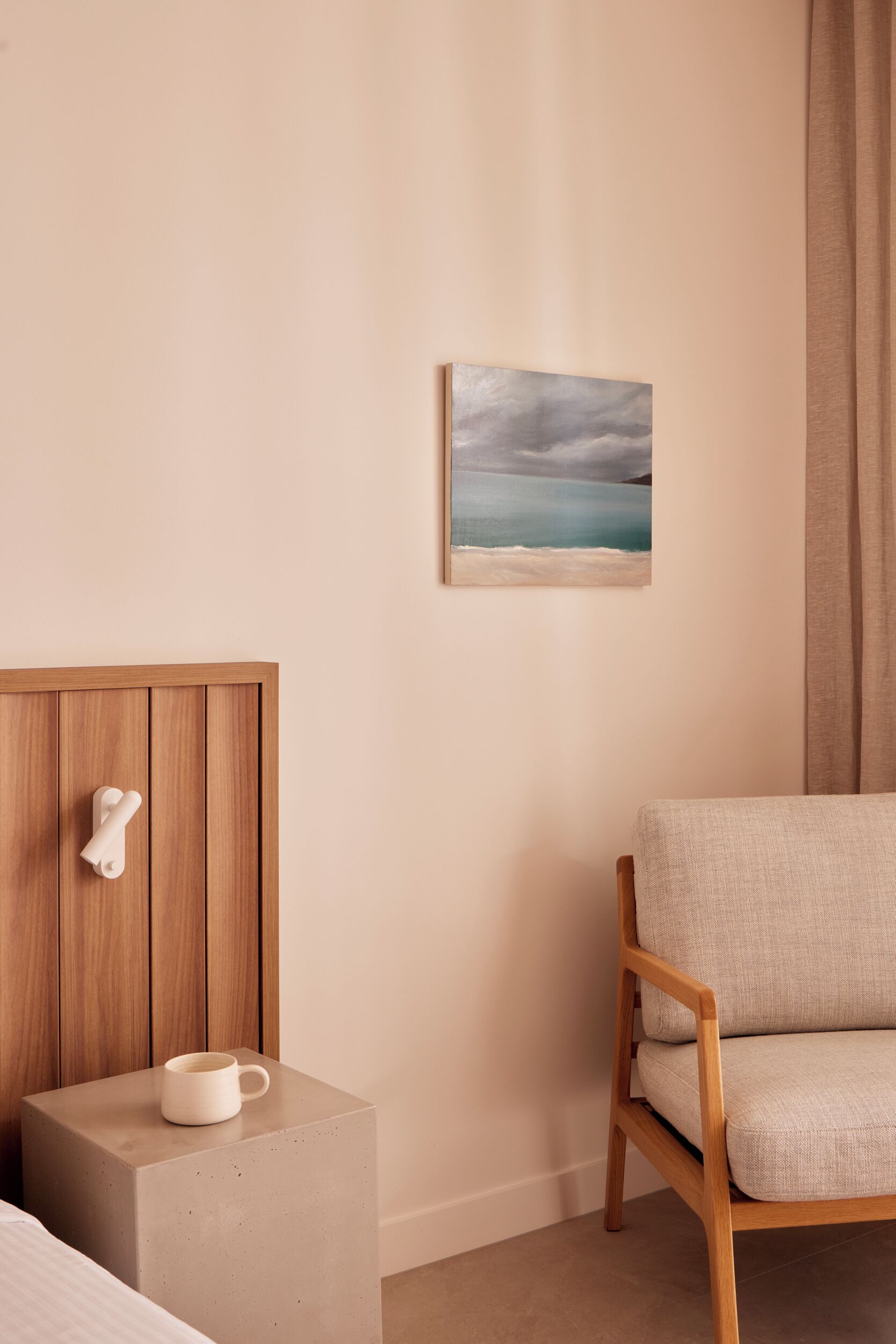A room with a wooden headboard, a white mug on a small side table, a beige sofa, and a painting of a coastal scene on the wall.