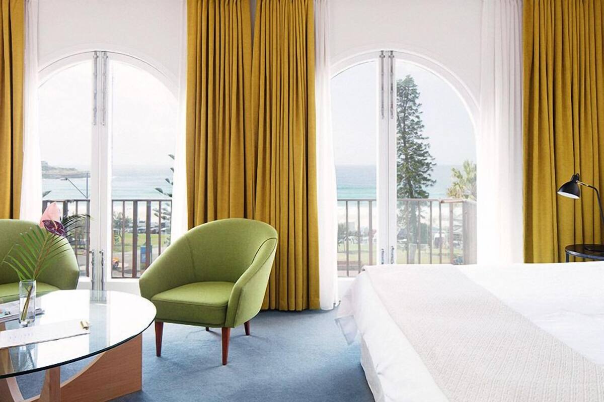 A hotel room with a bed, two green armchairs, a glass coffee table, and a view of the ocean.