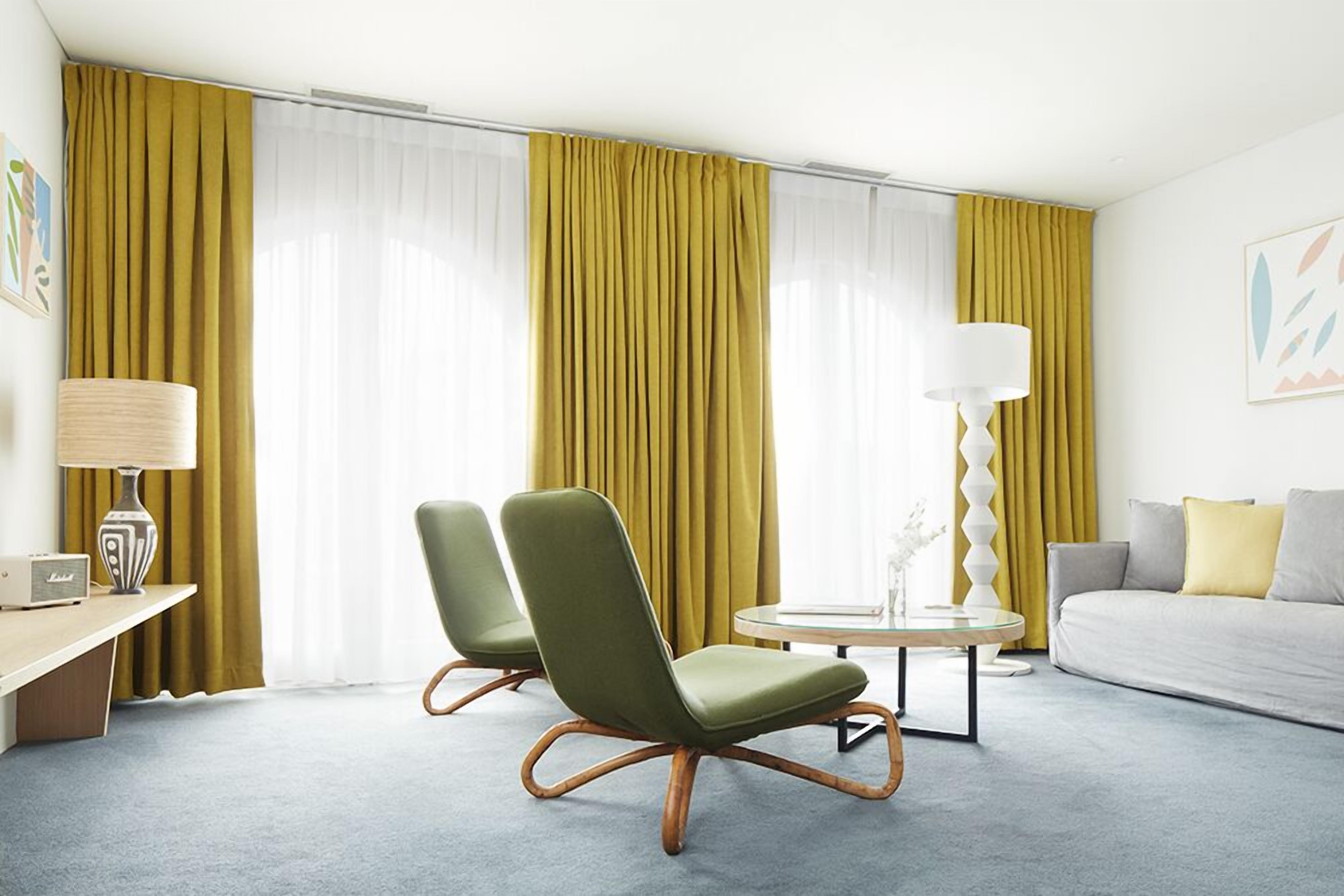 A modern hotel room with a sofa, two green armchairs, a round coffee table, a lamp, and a radio.