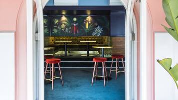 Una puerta abierta que da a un restaurante con alfombra azul, taburetes rojos y un mural de plantas tropicales.