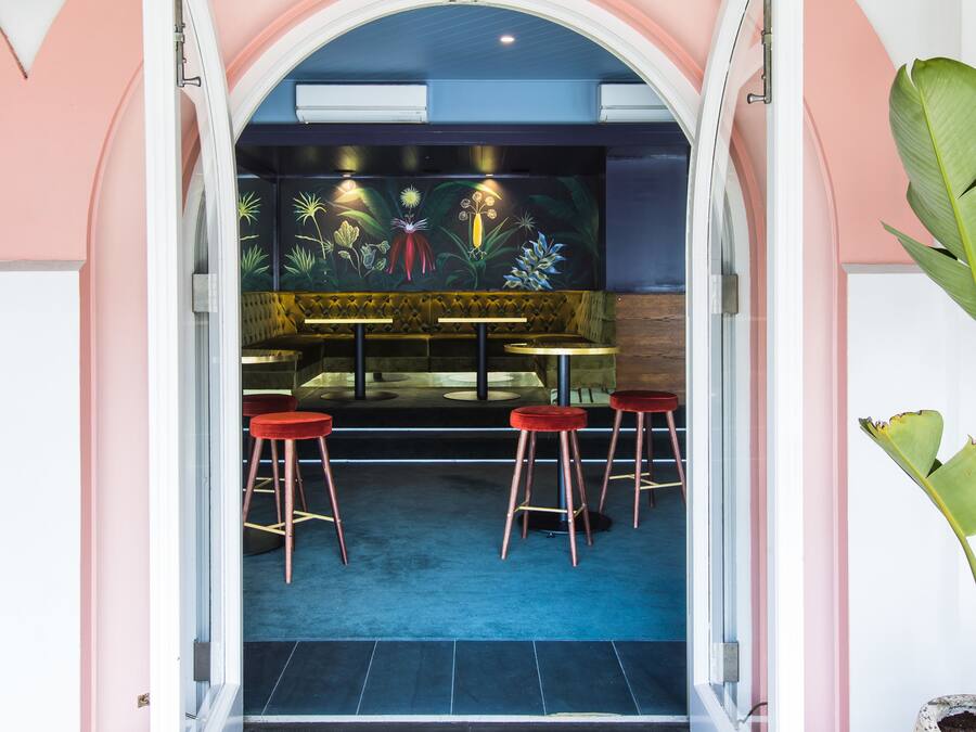 Una puerta abierta que da a un restaurante con alfombra azul, taburetes rojos y un mural de plantas tropicales.