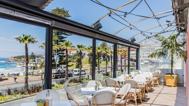 A terrace with wicker chairs and tables, overlooking a beach and cityscape. - Hotel Ravesis (Bondi Beach)