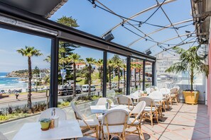 A terrace with wicker chairs and tables, overlooking a beach and cityscape.