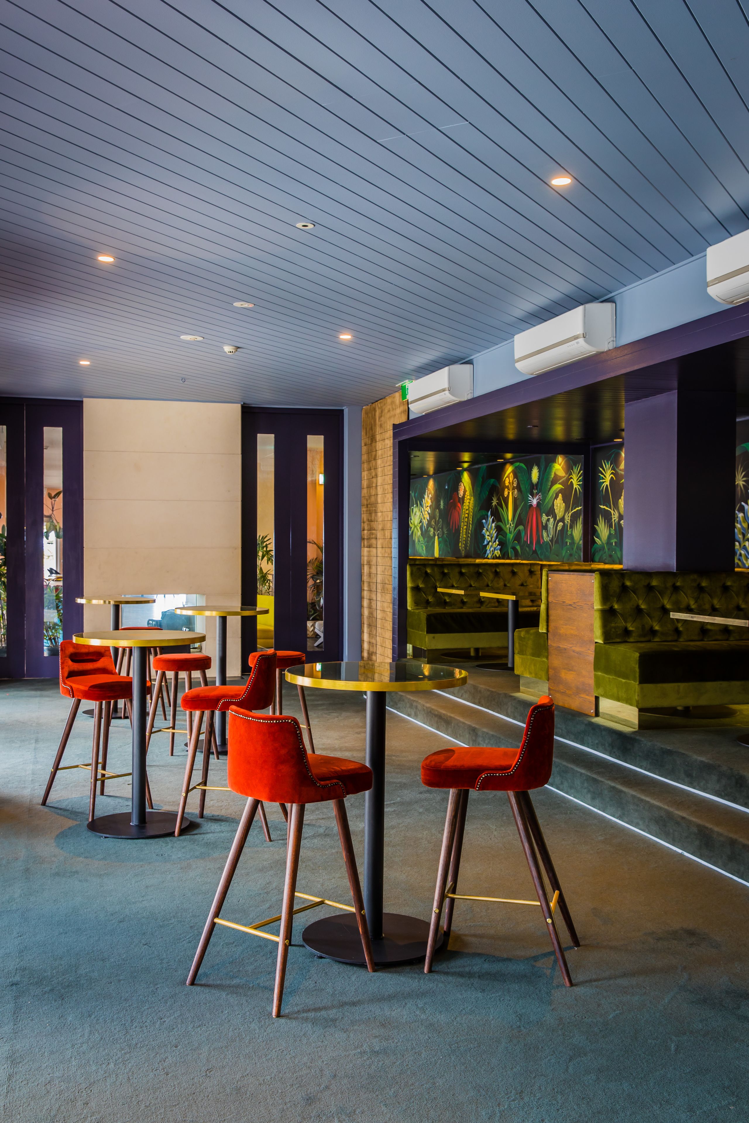 A modern indoor space with round tables and red chairs, featuring a ceiling with recessed lighting and a mural of tropical plants on the wall.