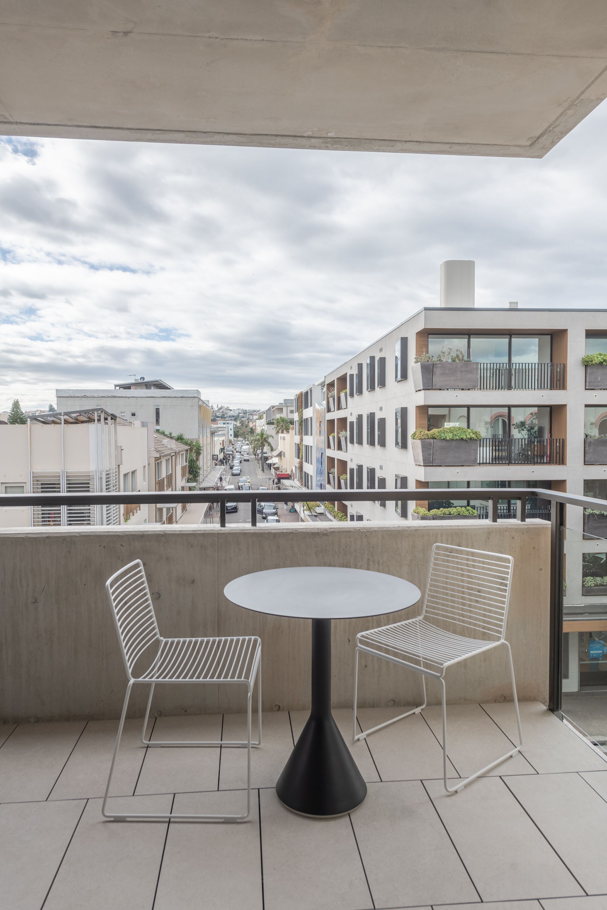 A balcony with two white wire chairs and a round table, overlooking a street with buildings and parked cars.