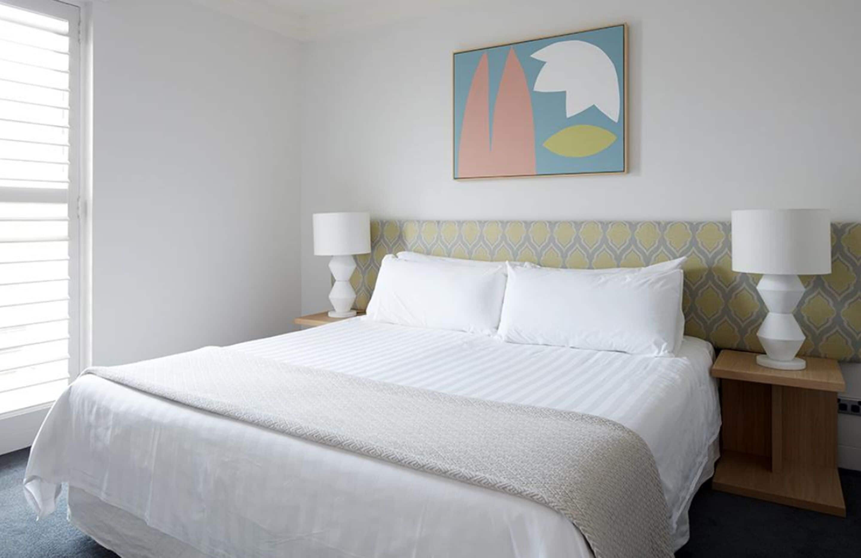 A hotel room with a large bed, two bedside lamps, a wall-mounted abstract painting, and a window with blinds.