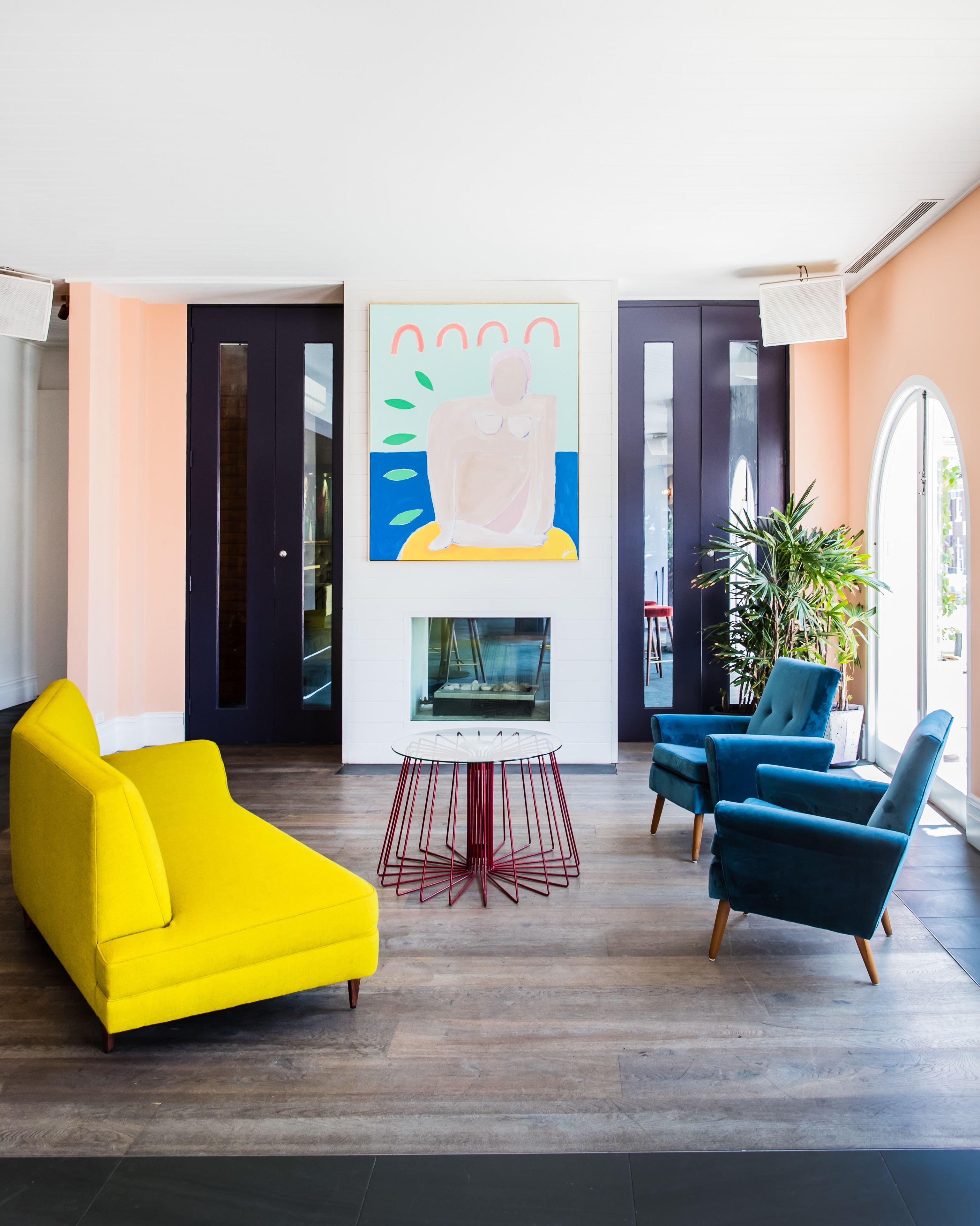 Una sala de estar moderna con un sofá amarillo, sillones azules y una mesa central. Hay un cuadro grande en la pared y una planta en maceta en la esquina.