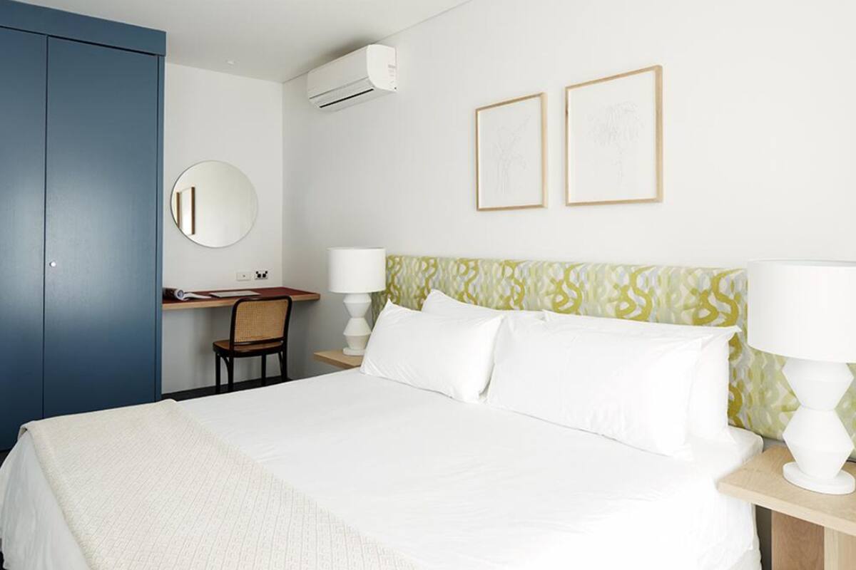 A hotel room with a bed, a desk, a chair, a round mirror, two framed pictures, and an air conditioning unit.