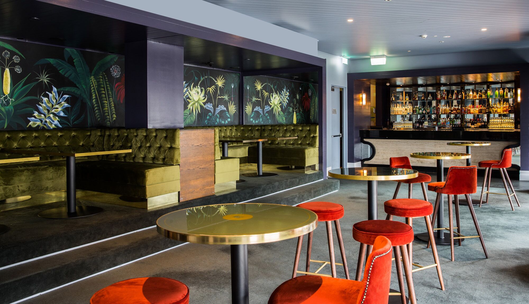 A bar area with high stools and a bar counter, featuring a mural of tropical plants on the wall.