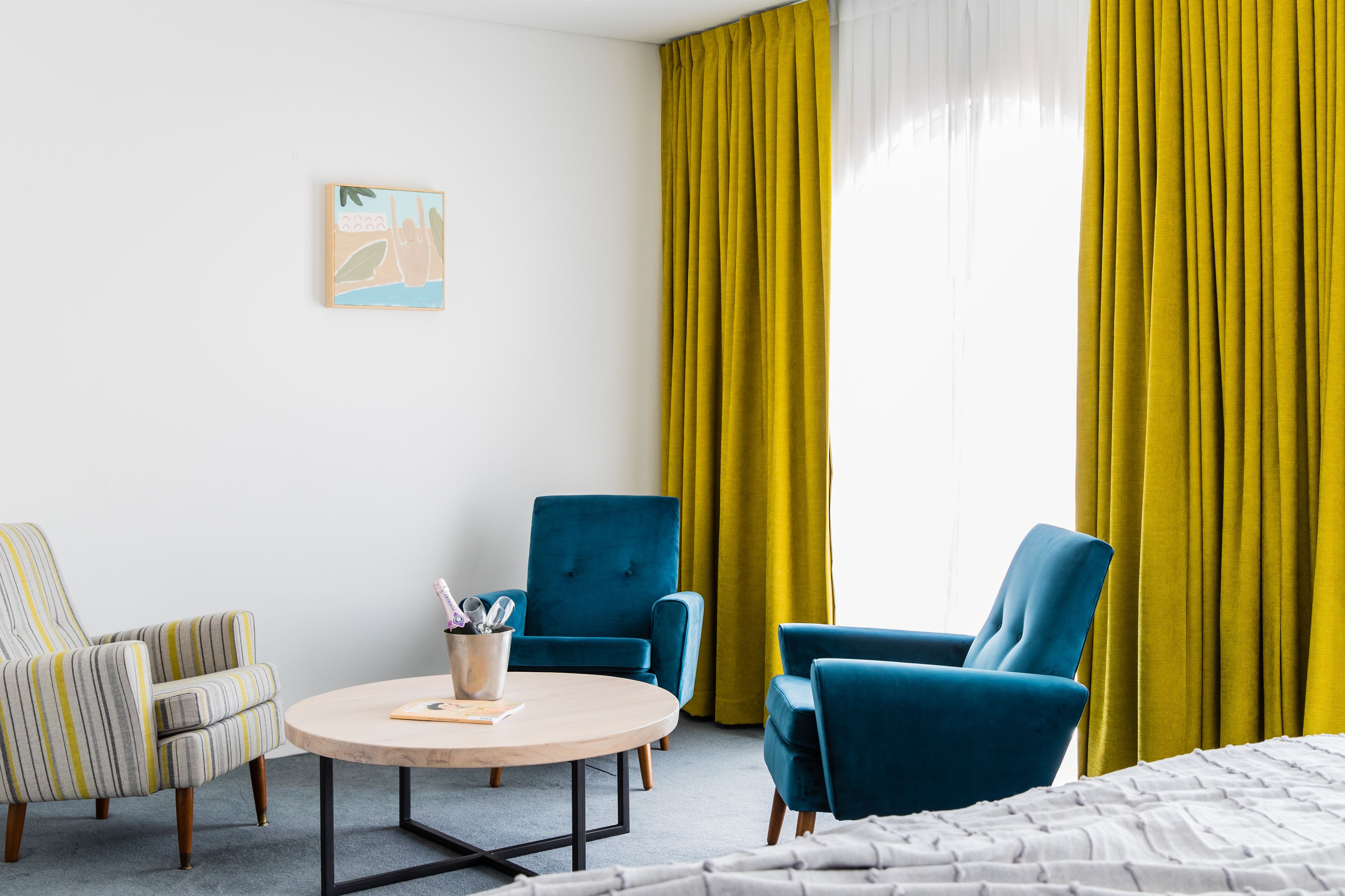A hotel room with a bed, a round table with a book and a pen holder, two blue armchairs, a striped armchair, and a yellow curtain.