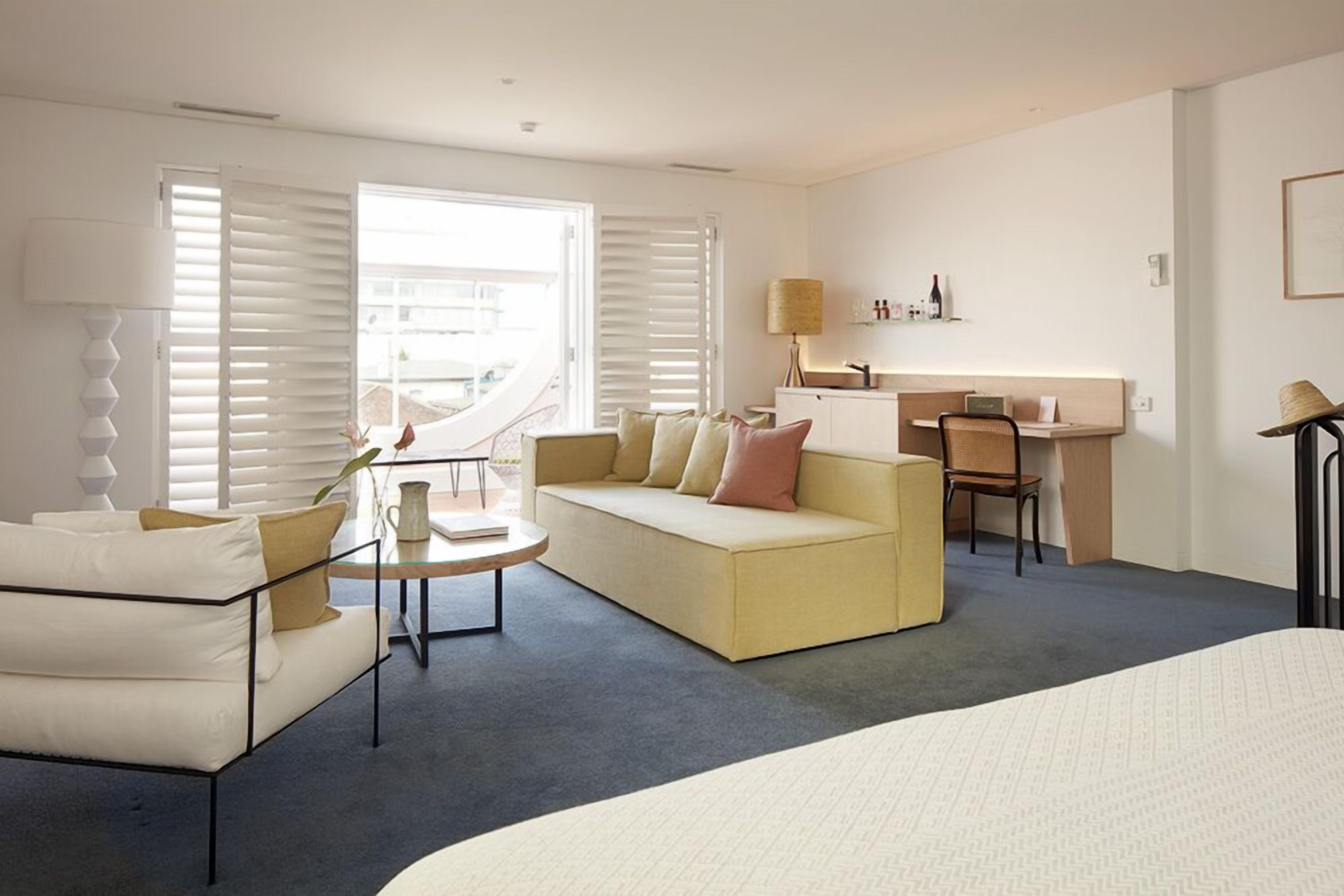 A modern hotel room with a sofa, a small round table, a desk with a chair, and a bed.