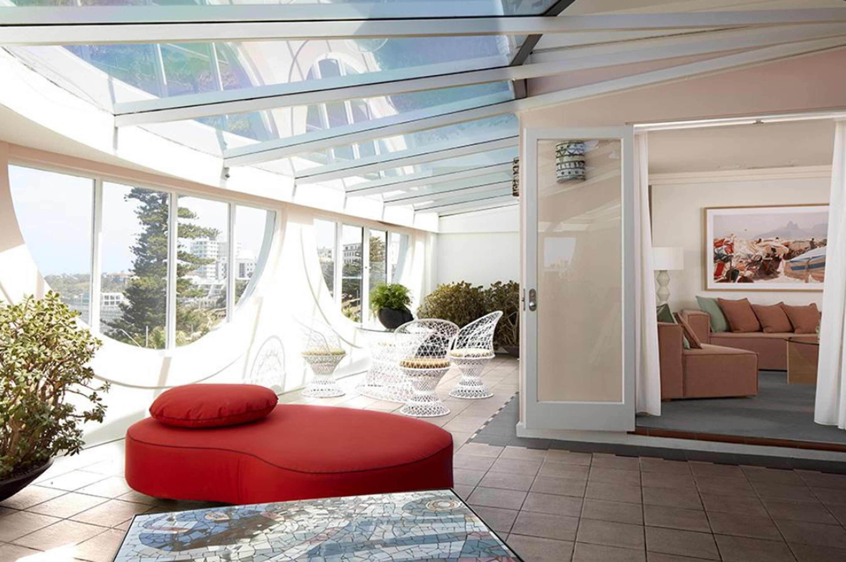 Un interior moderno con techo de vidrio, un área de descanso con cojines rojos, muebles de mimbre blanco y vistas a la ciudad.