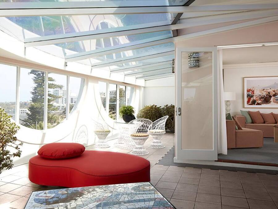 Un interior moderno con techo de vidrio, un área de descanso con cojines rojos, muebles de mimbre blanco y vistas a la ciudad.