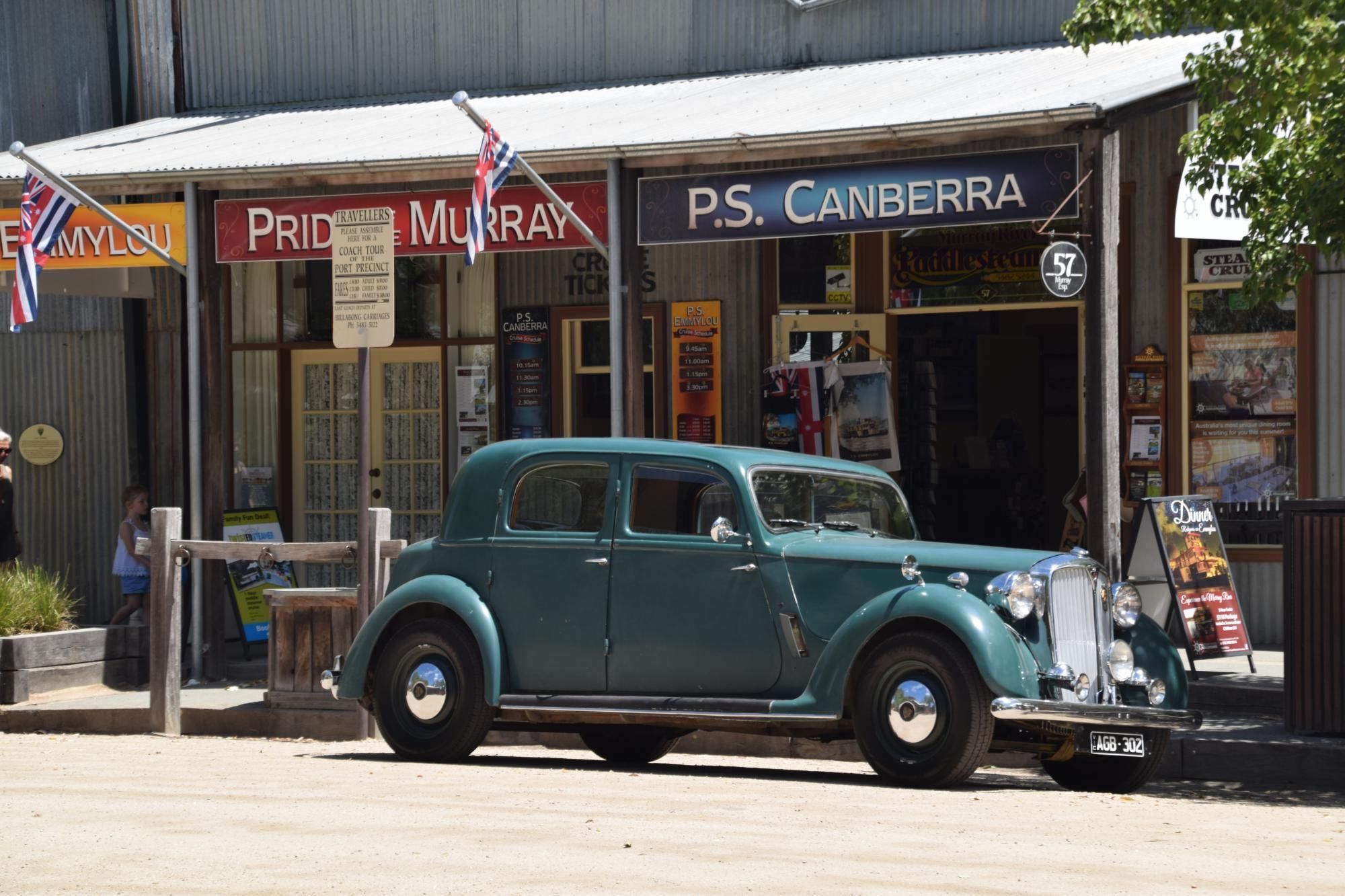 Une voiture ancienne de couleur turquoise est garée devant un bâtiment où l’on peut voir des enseignes indiquant « Pride Murray » et « P.S. Canberra ».