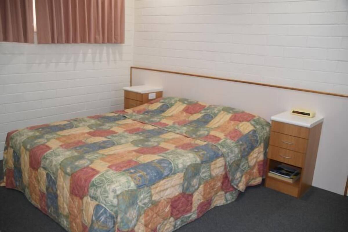 A single bed with a colorful quilt, a bedside table with a book, and a small drawer unit.