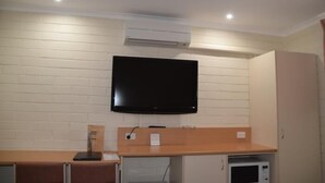A room with a flat-screen TV mounted on a white brick wall, a wooden counter with a microwave below, and a white air conditioning unit on the ceiling. - Big River Motel (Echuca)