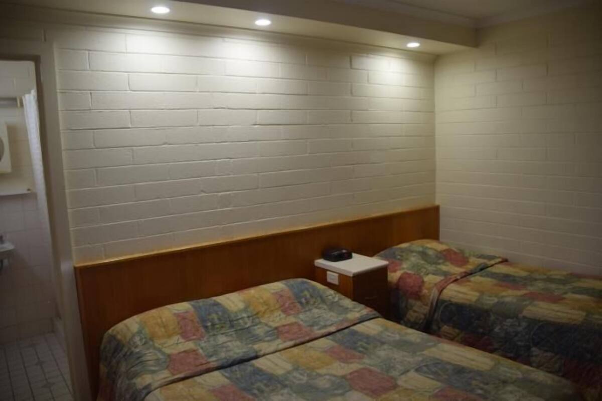 A small, compact room with two single beds, a wooden headboard, a small bedside table, and a white brick wall.