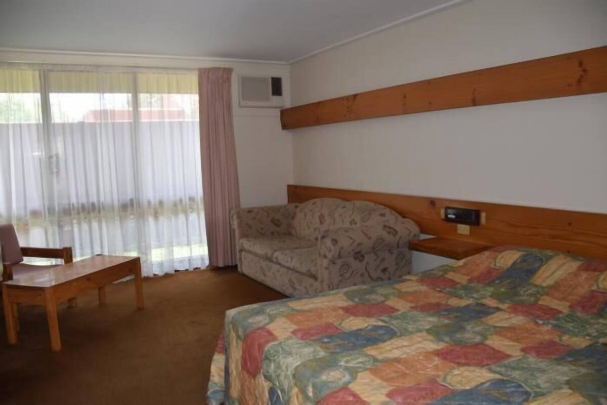 A room with a bed, a wooden headboard, a sofa, a table, and a window with curtains.