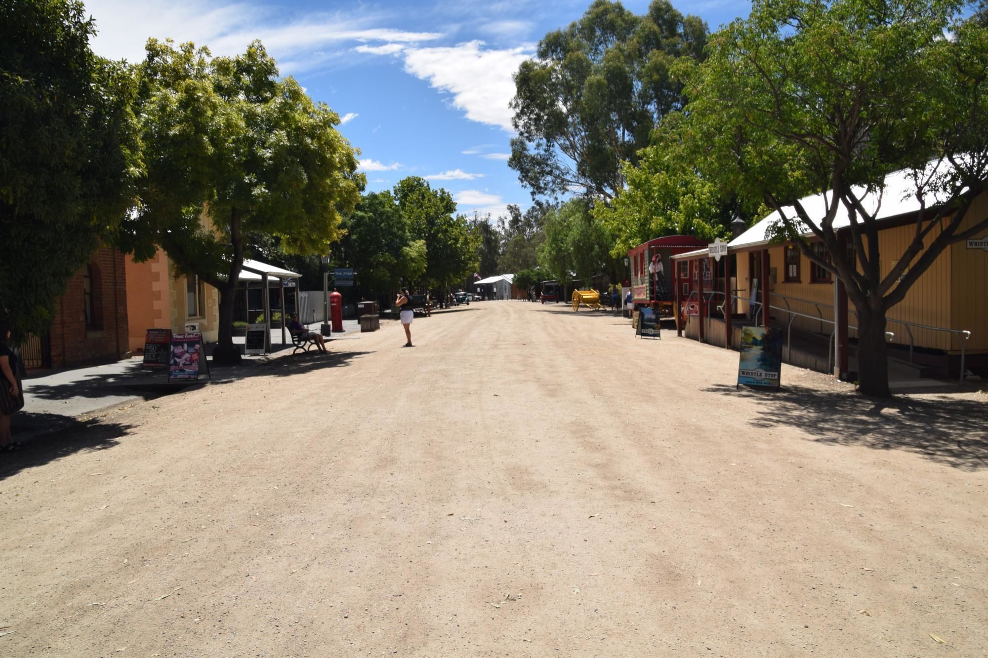 Una calle rural bordeada de árboles y edificios, incluyendo una tienda y una parada de autobús.