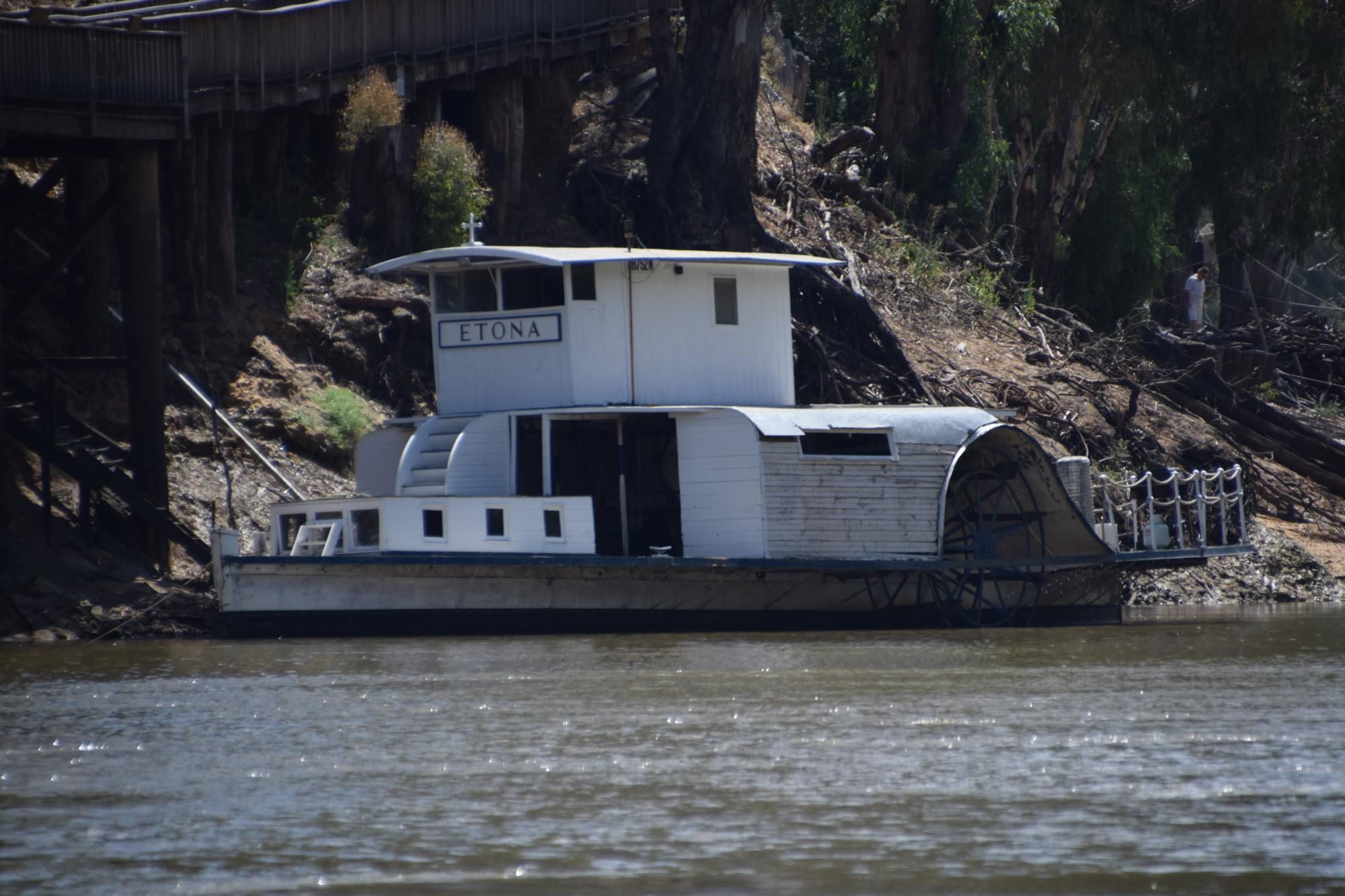 A white houseboat with a covered deck and a small cabin labeled "ETONA" on a river.