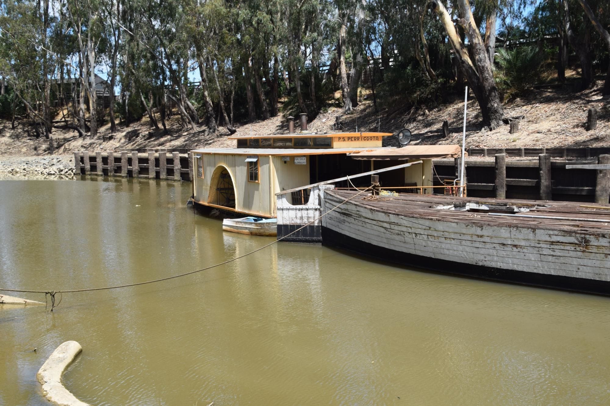 A houseboat docked at a river with a wooden deck and a small boat in front.