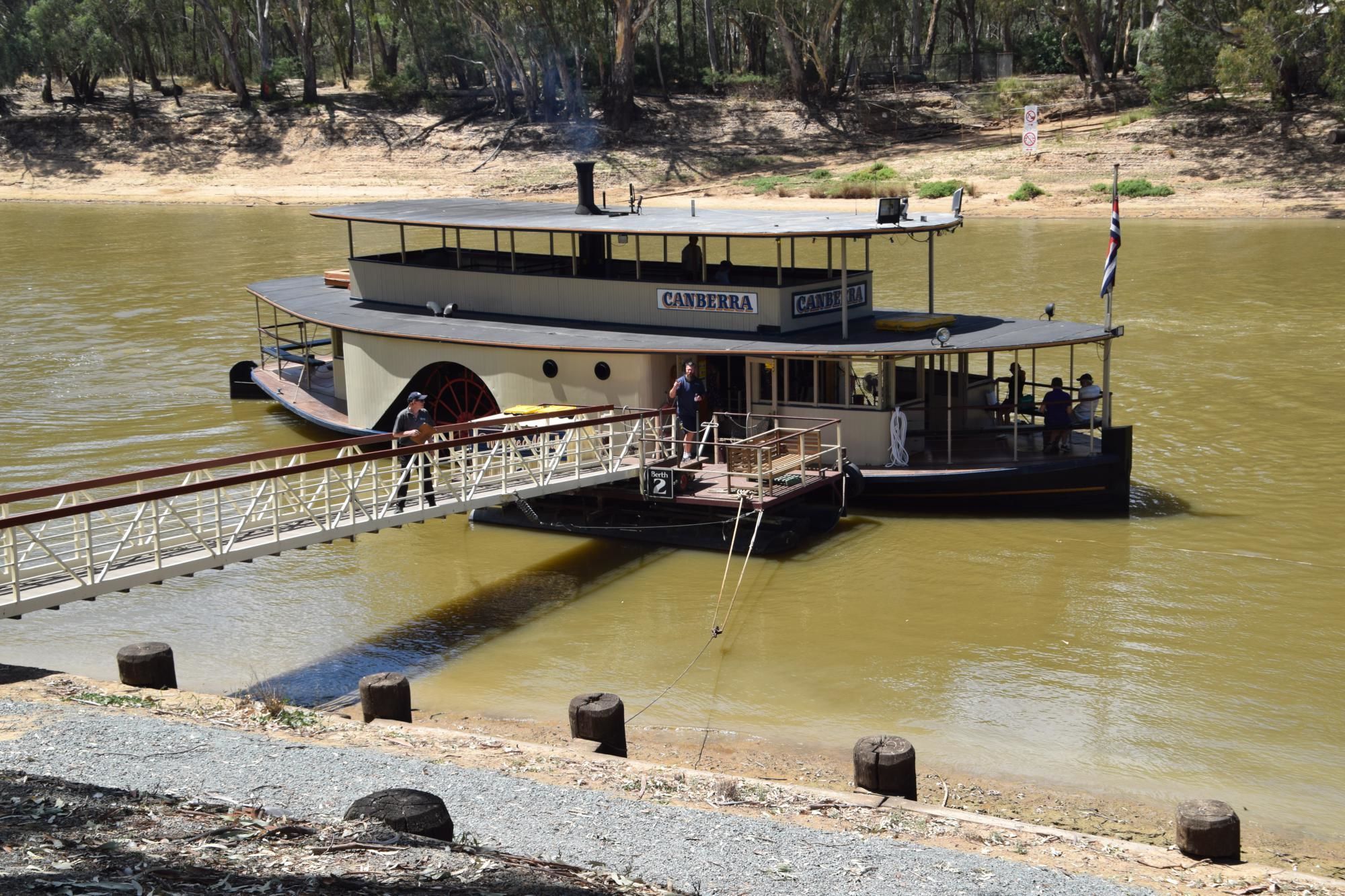 A riverboat with a covered upper deck and a lower open deck, docked at a pier.