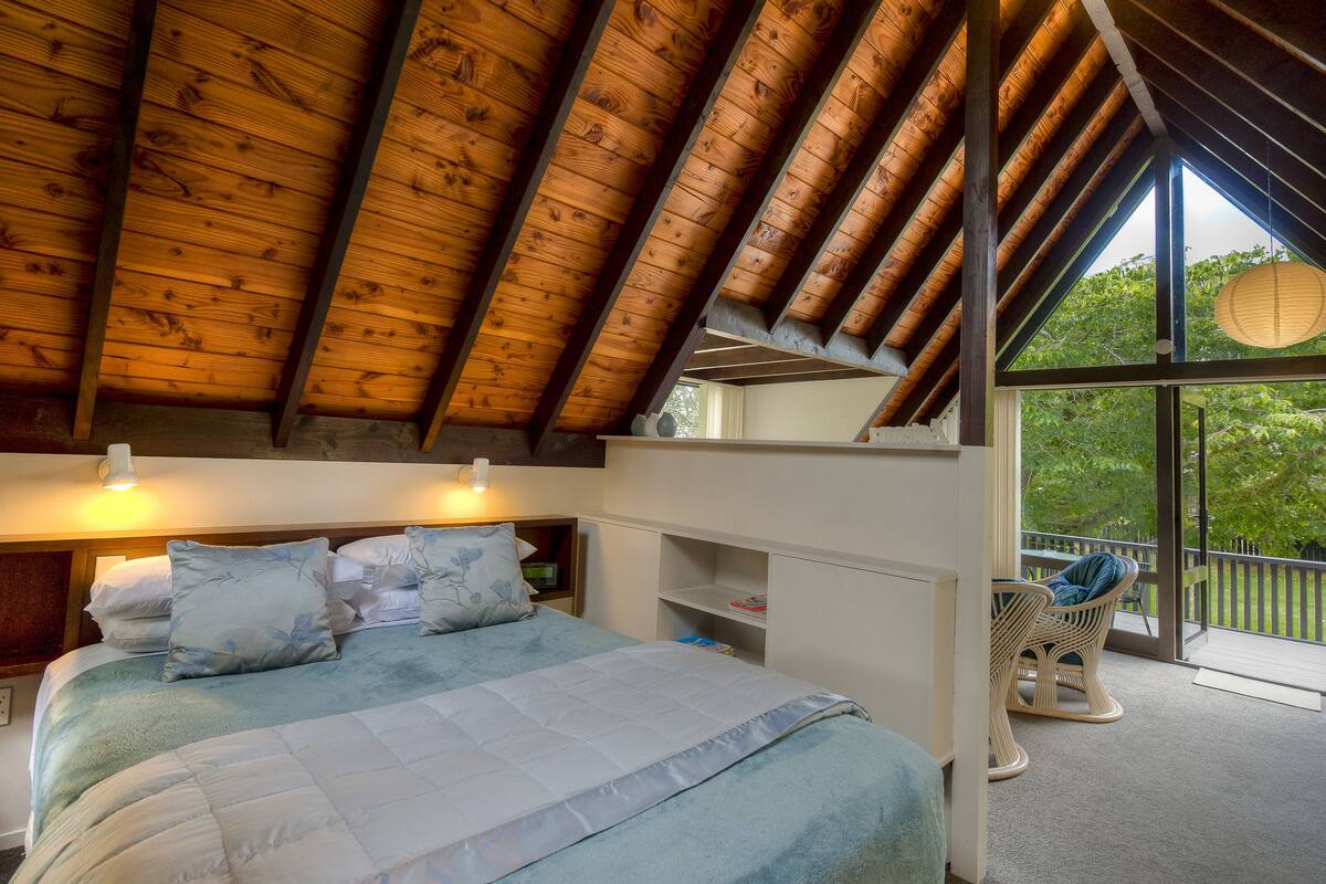 A bedroom with a sloped ceiling, a bed with a blue quilt, and a view of the outdoors through large windows.