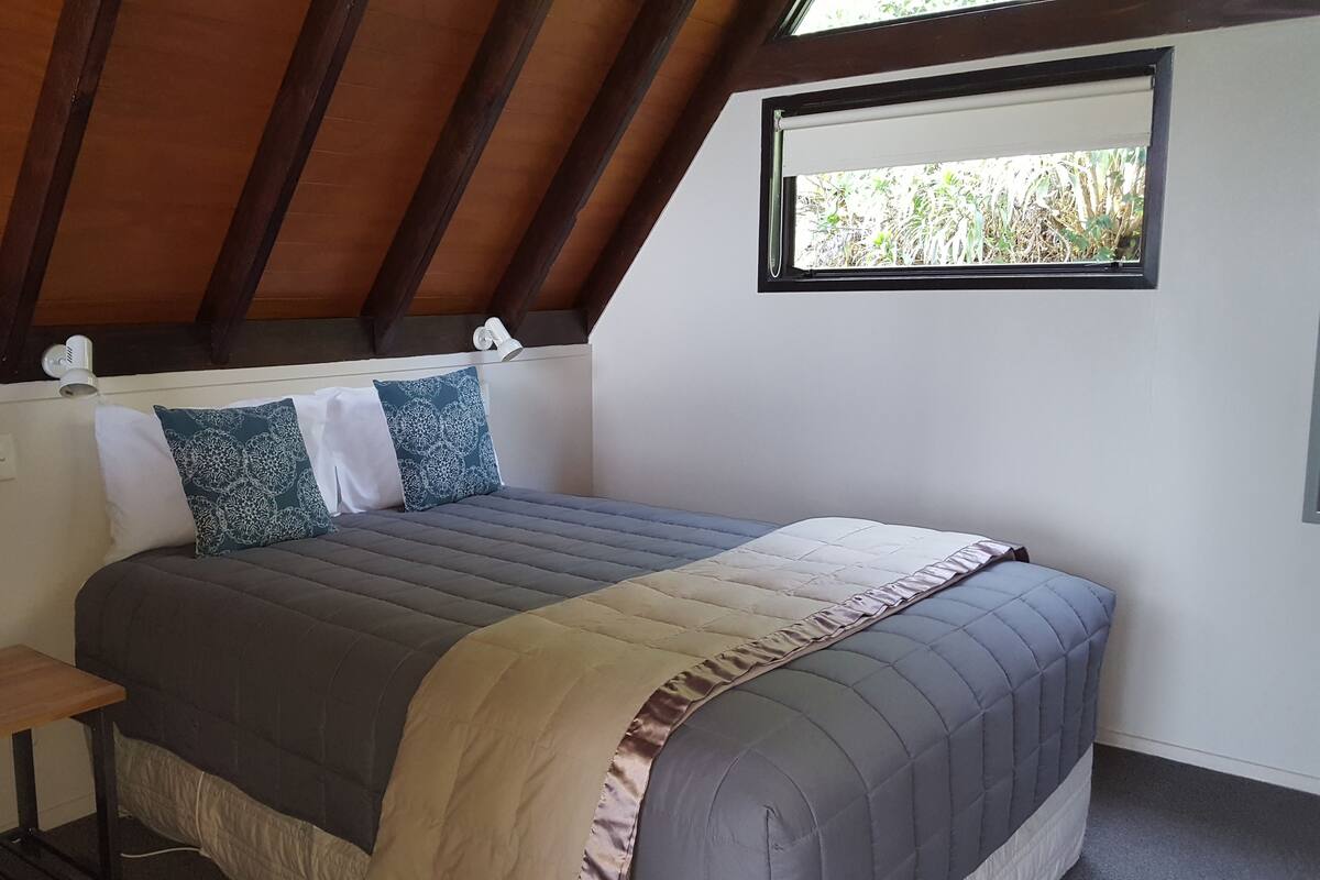 A bedroom with a bed, pillows, a bedside table, a window, and a ceiling with wooden beams.