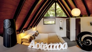 A cozy attic bedroom with a sloped ceiling, a bed with a white headboard, a small window, and a door.