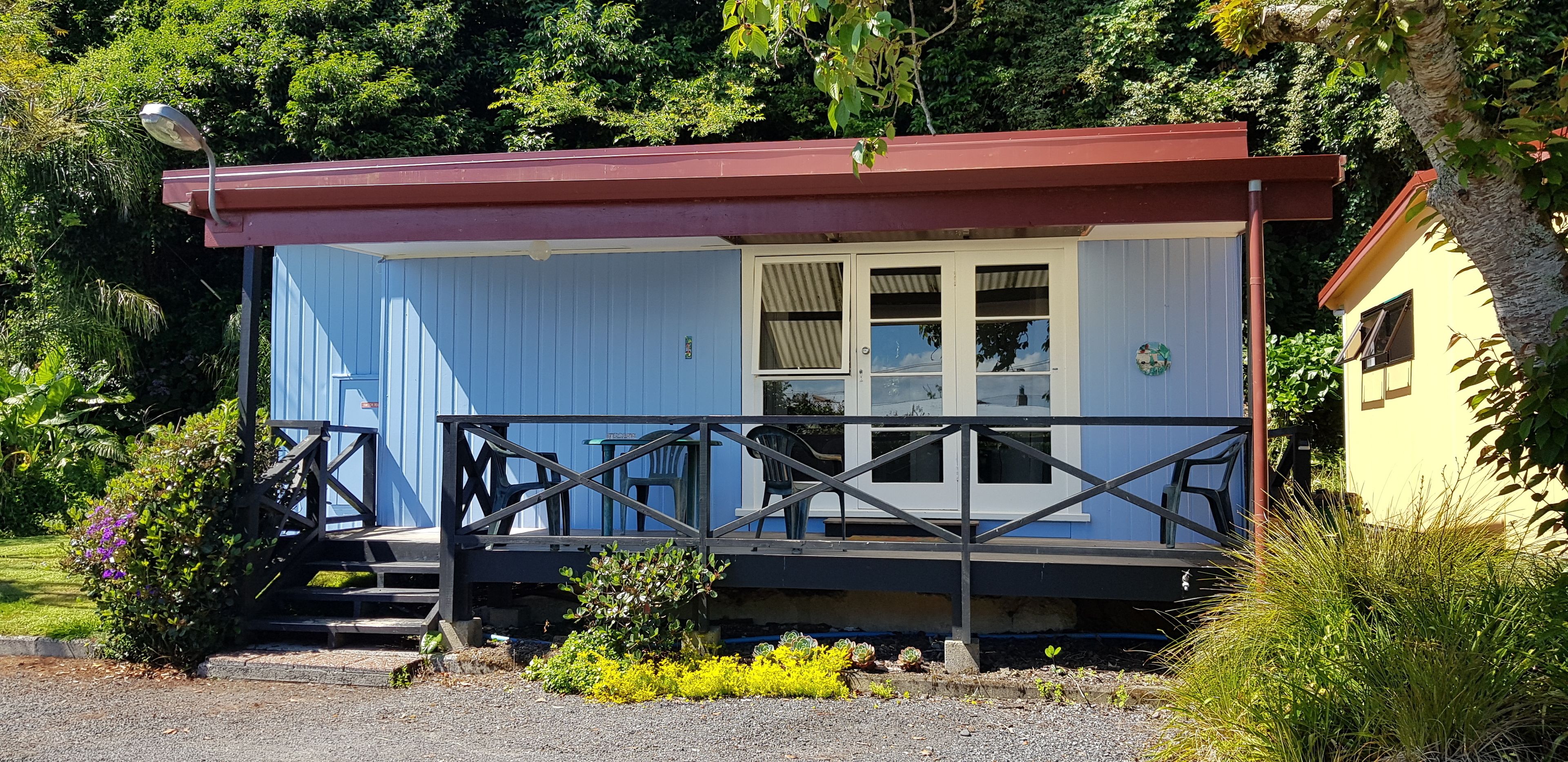 A small, single-story building with a blue exterior and a red roof. It features a porch with a railing and several chairs.