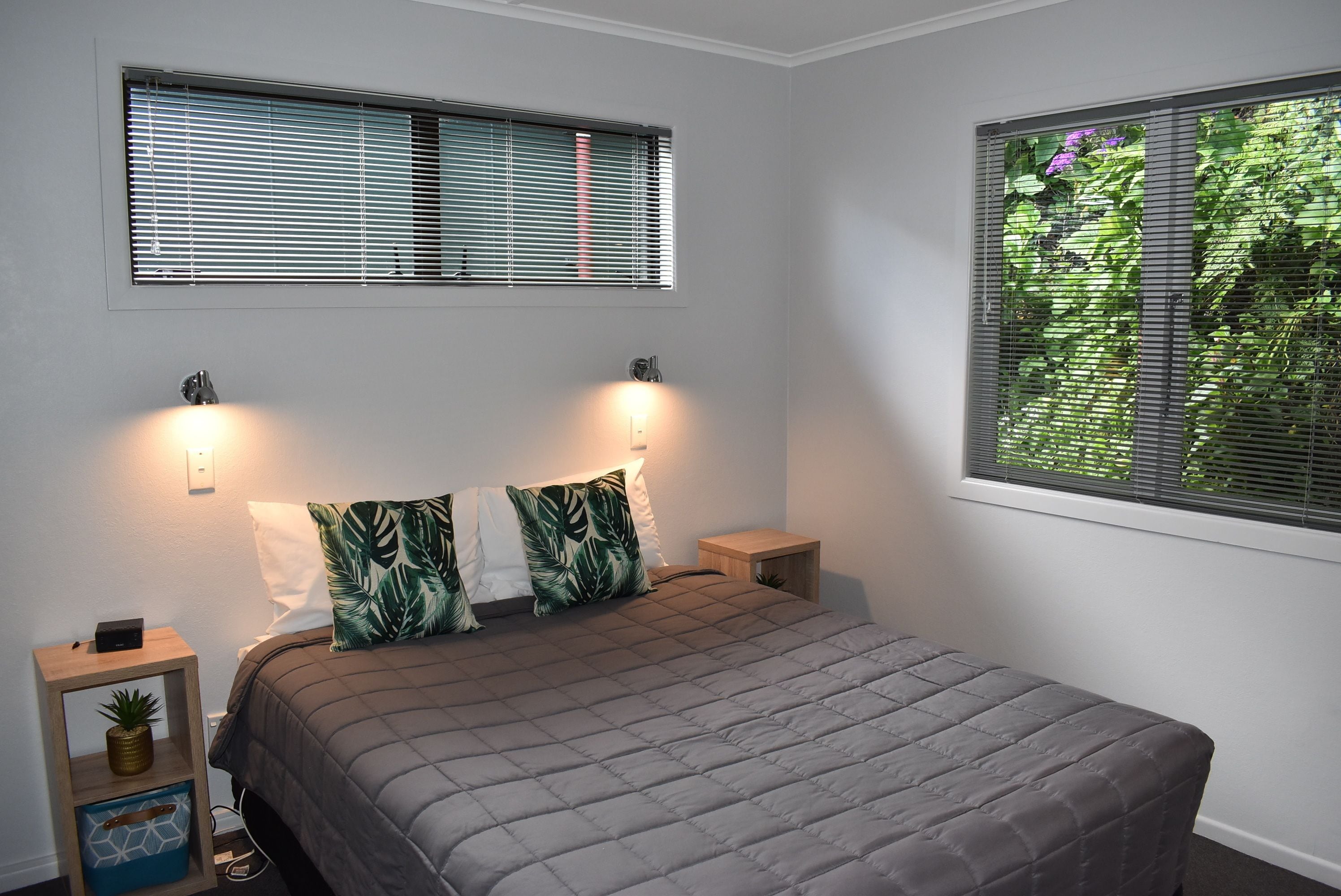 A bedroom with a bed, two bedside lamps, a nightstand with a plant, and a window with blinds.