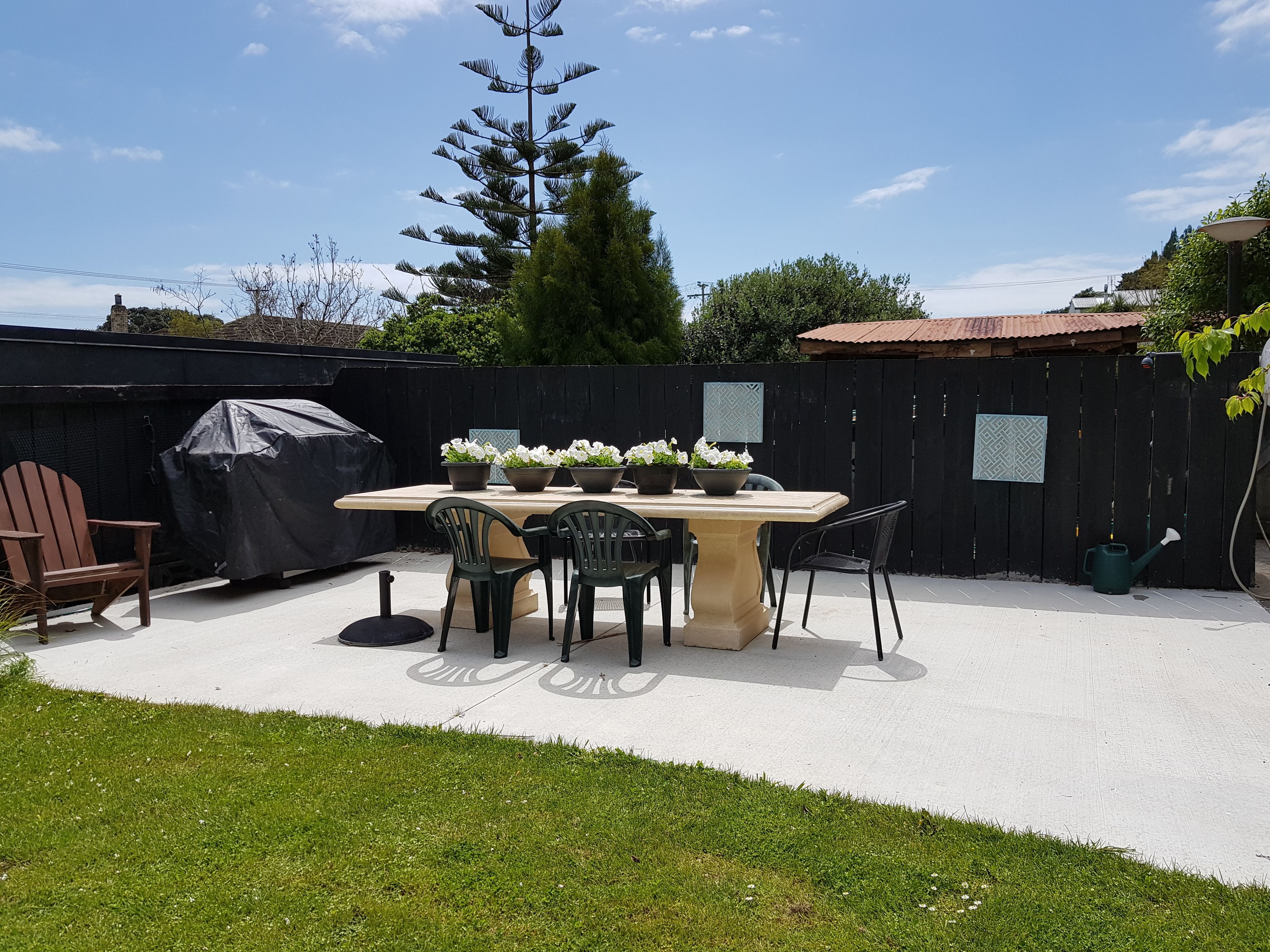 A patio with a table and chairs, surrounded by a black fence, with a tree and a covered grill in the background.