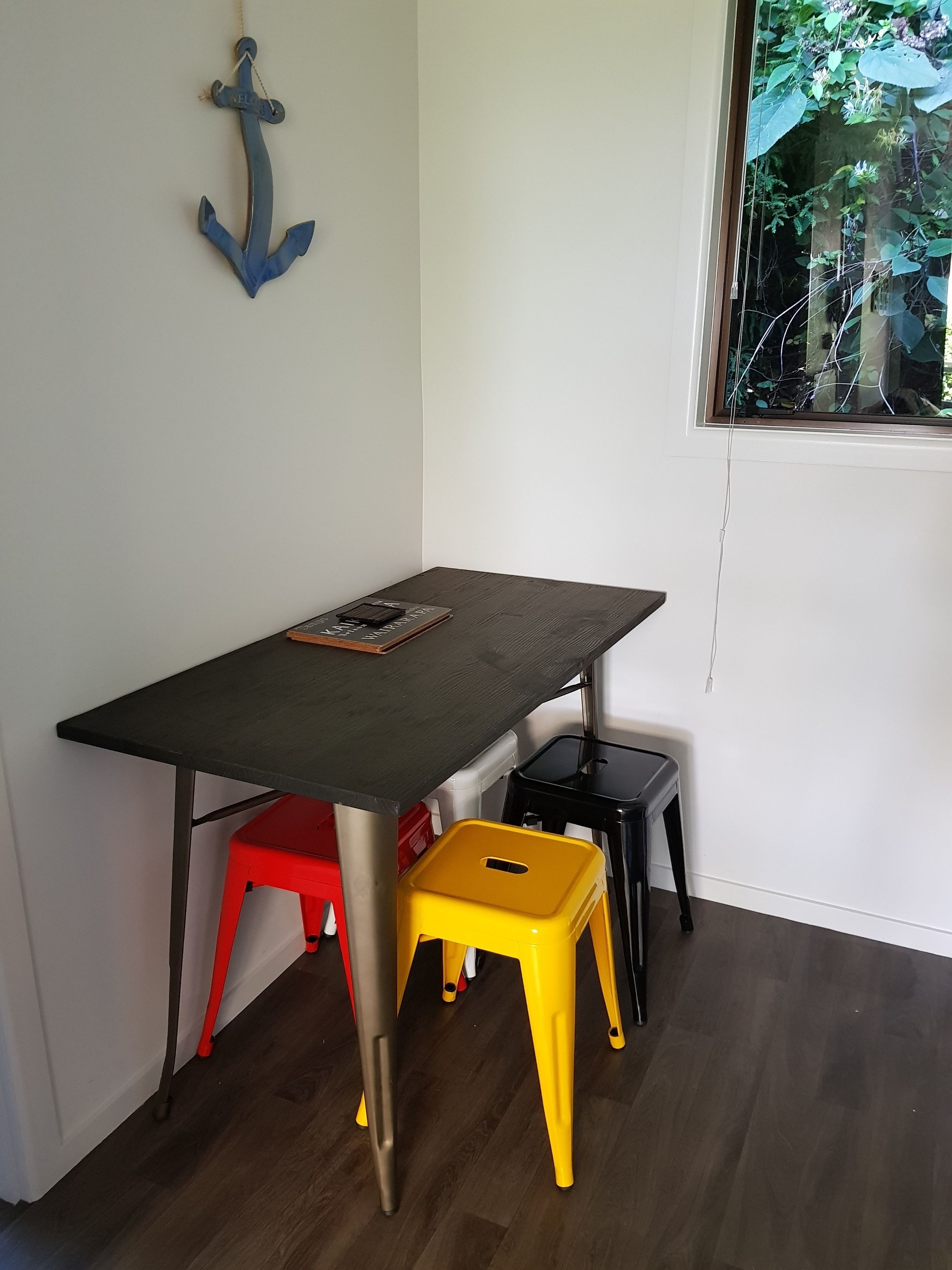 A corner room with a wooden table, a red and a yellow stool, and a blue anchor decoration on the wall.