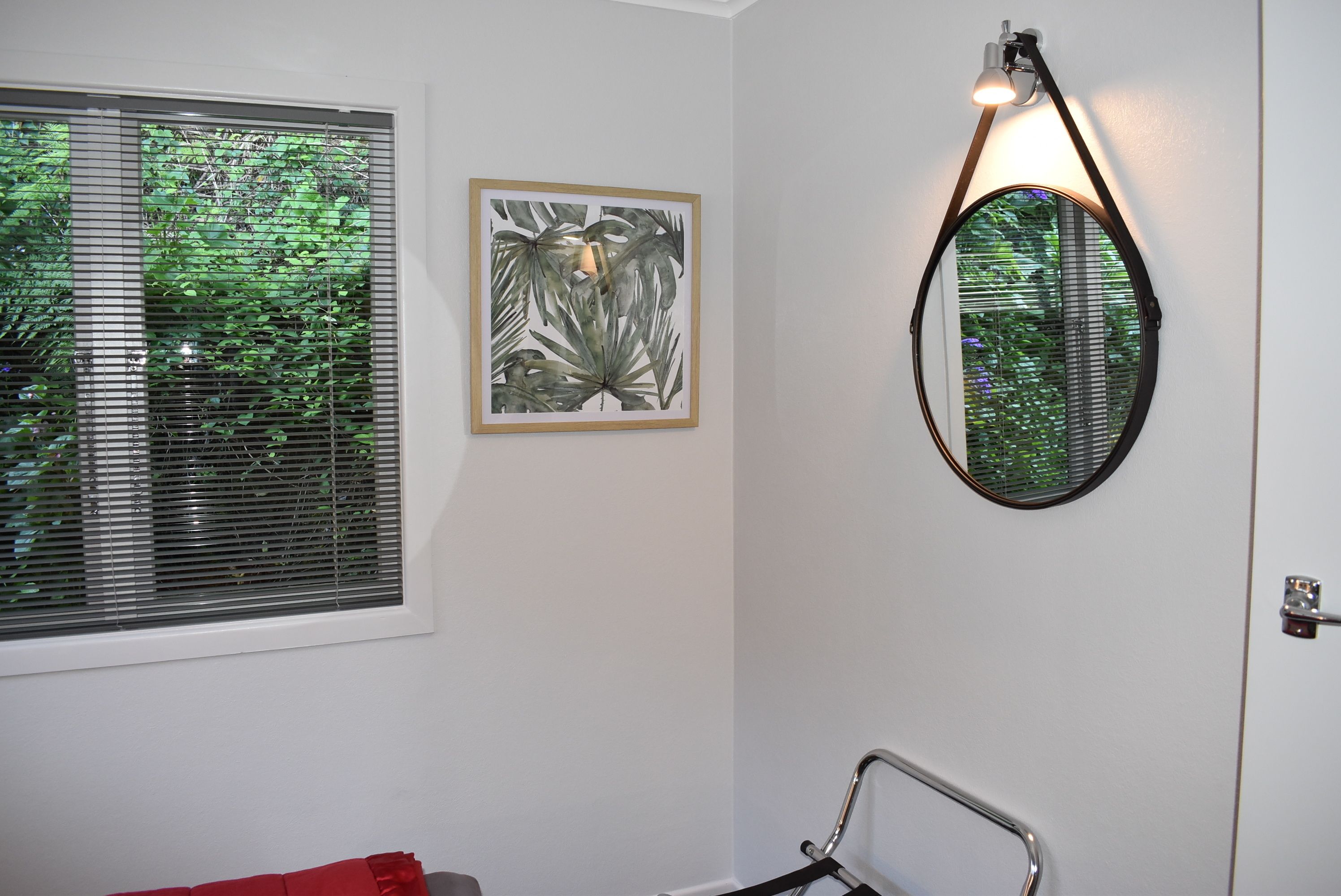 A bathroom with a window, a framed artwork, a round mirror, and a shower chair.