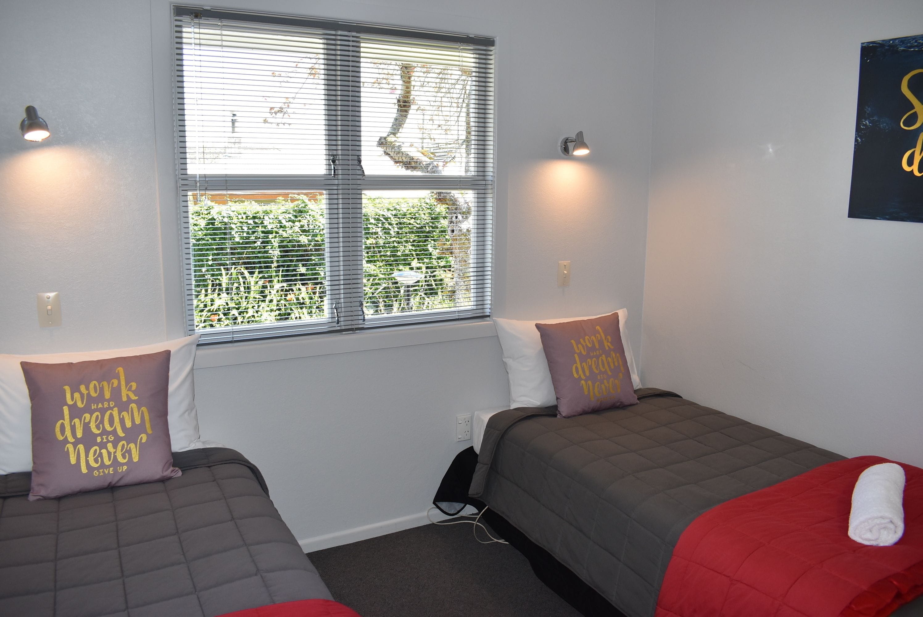 A room with two beds, each with a grey and red blanket, and a window with blinds.