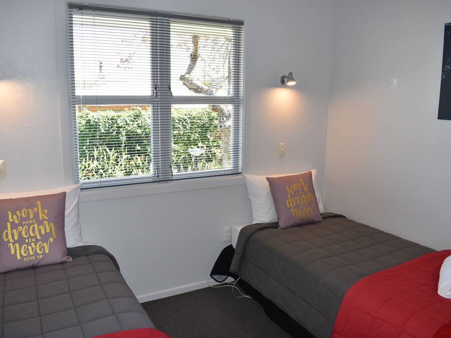 A room with two beds, each with a grey and red blanket, and a window with blinds.