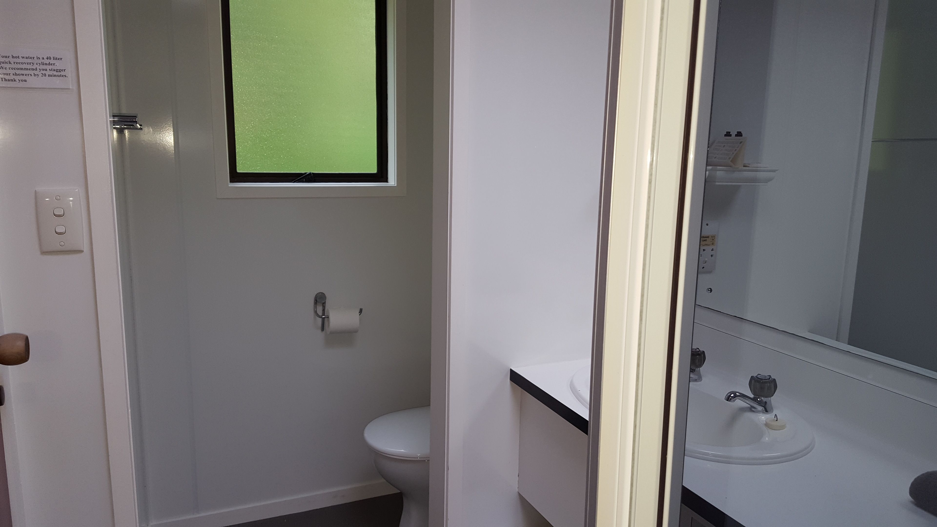 A bathroom with a toilet, a sink, and a mirror. There is a window above the toilet, and a sign on the wall.