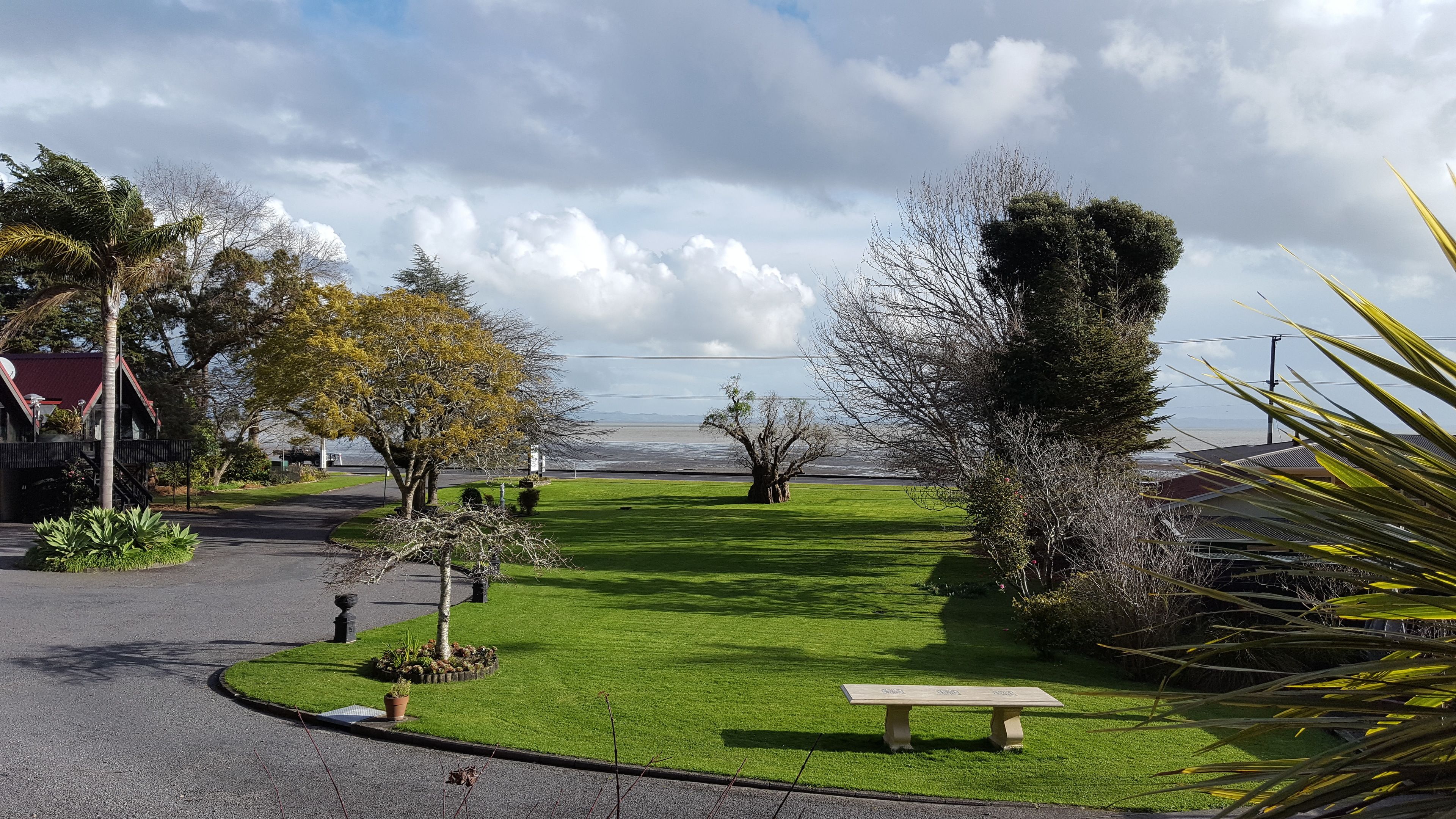 A well-maintained garden with a bench, various trees, and a clear view of the sea in the background.