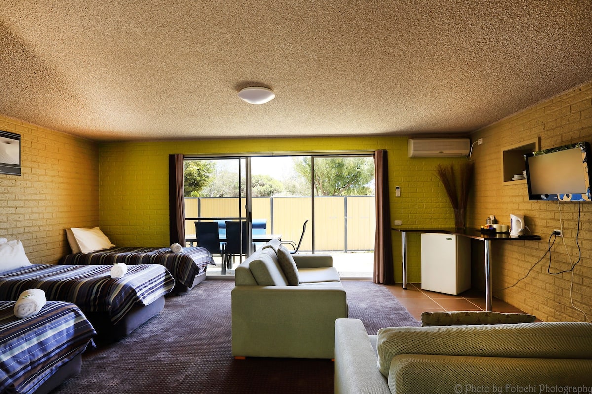 A room with two beds, a sofa, a small kitchenette, and a large window offering a view of the outdoors.