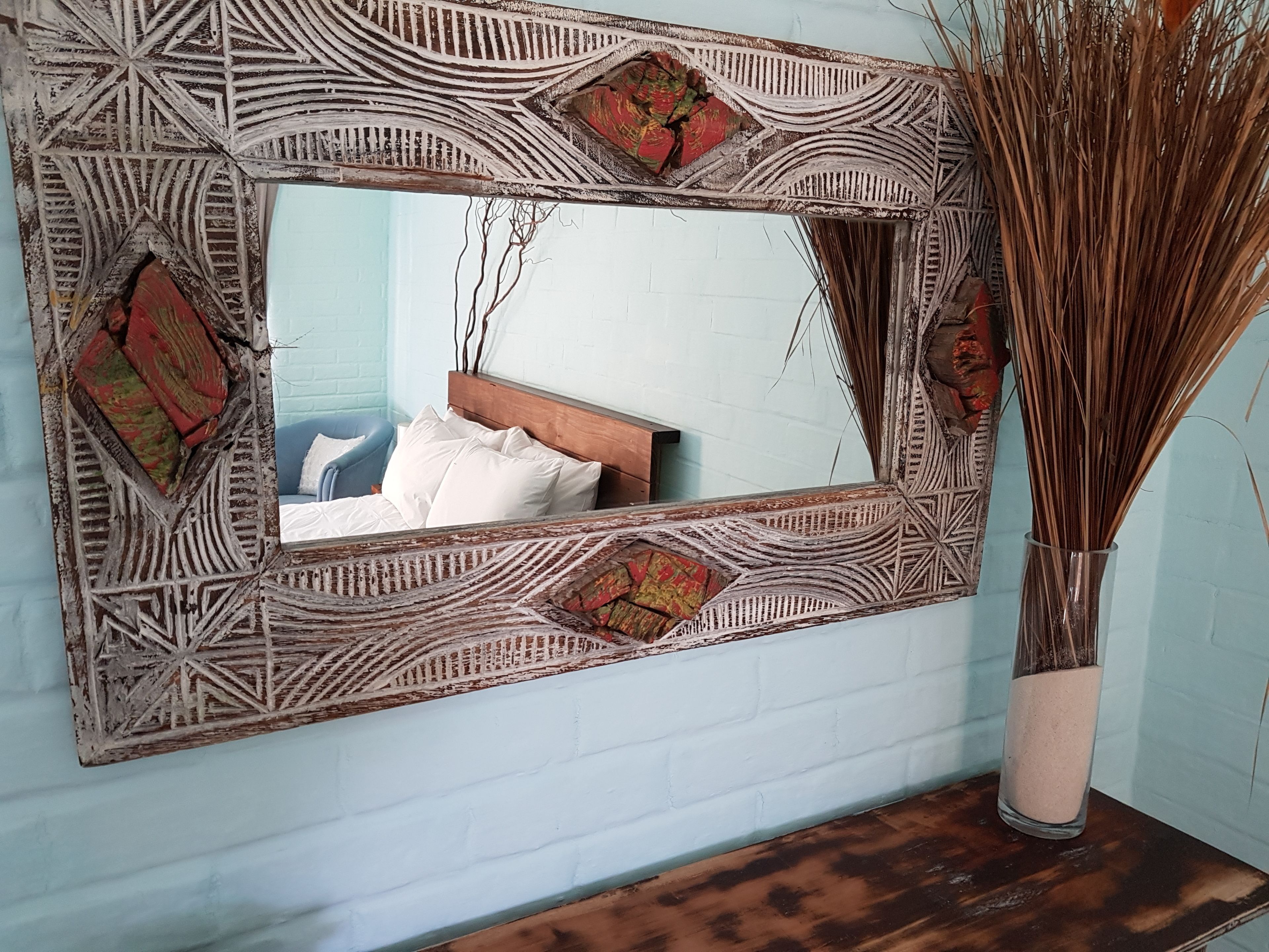 A mirror with intricate patterns and a bed with white pillows and a blue chair in the reflection.