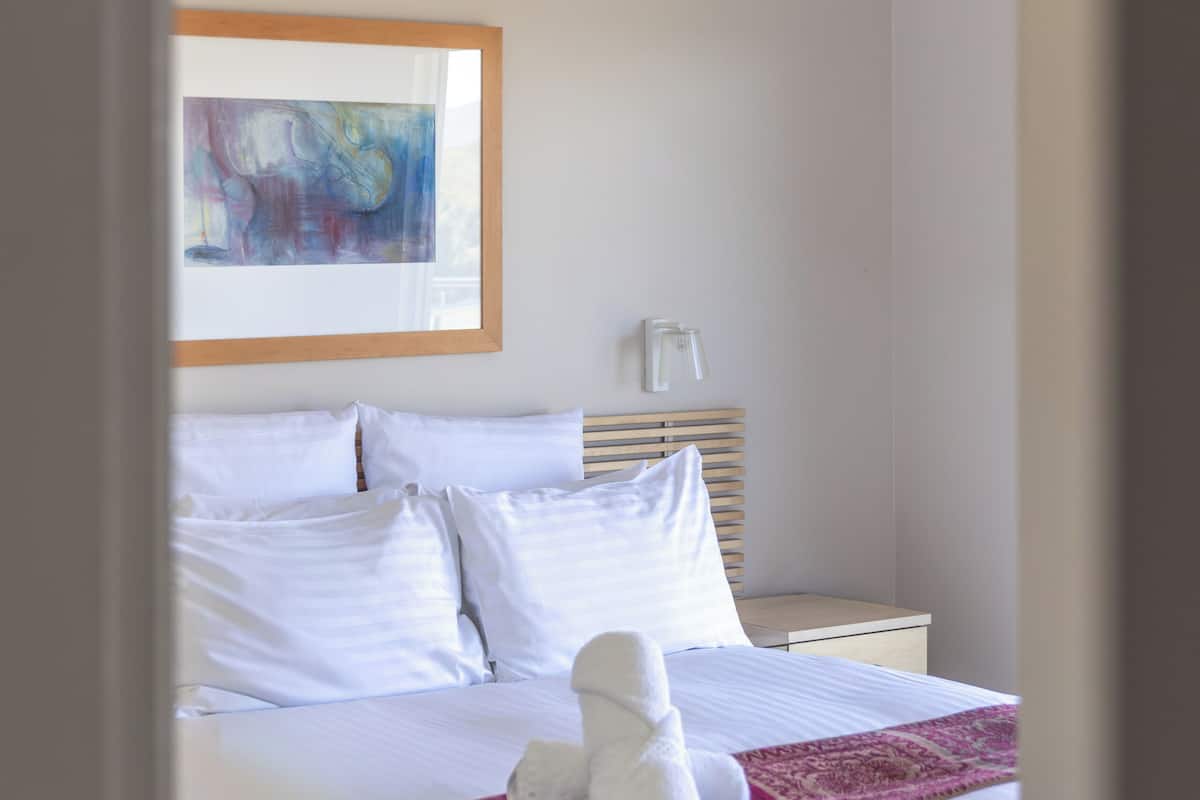 Una cama bien hecha con sábanas blancas, una manta roja y una toalla blanca.