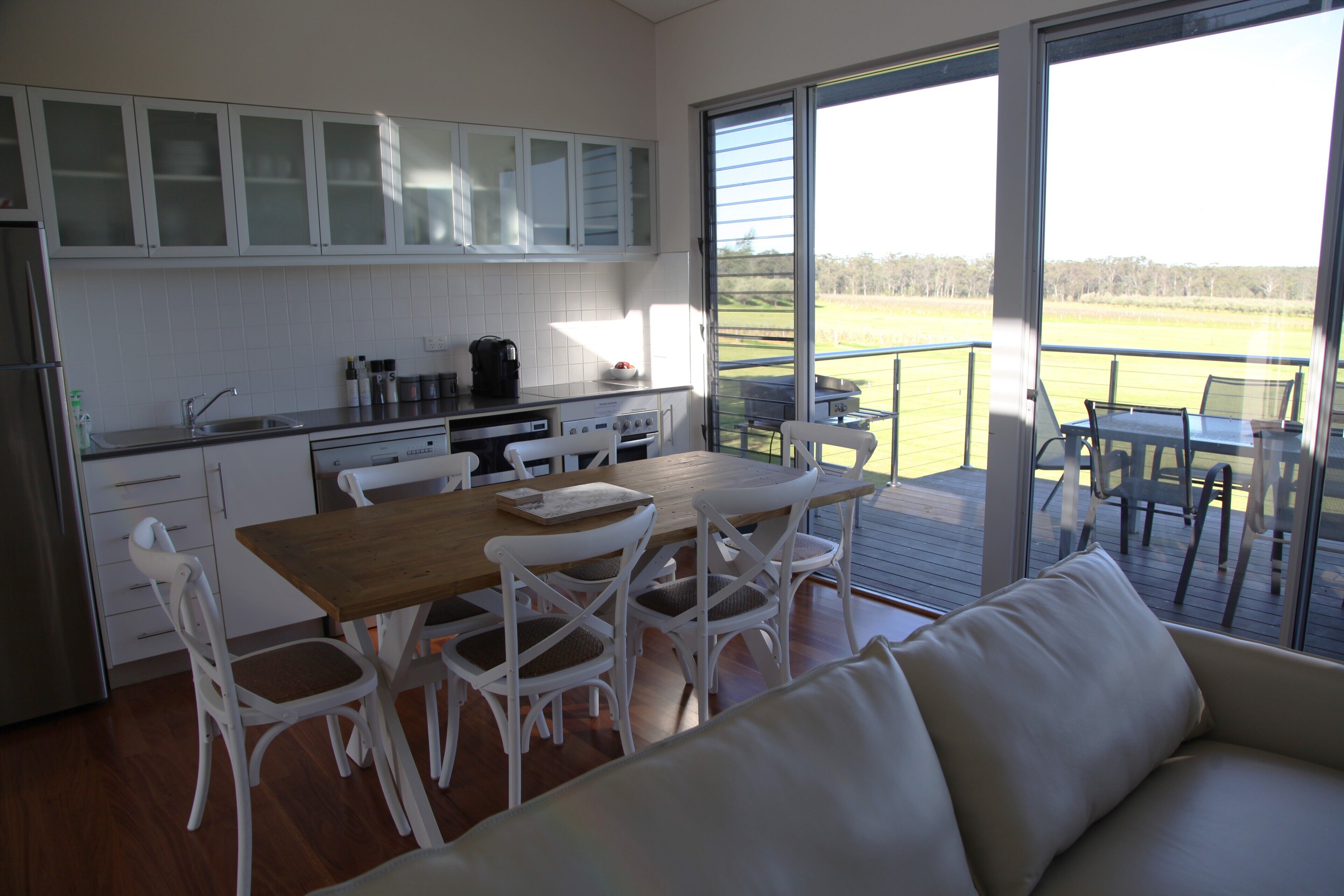 A modern kitchen with white cabinets, a wooden dining table, and white chairs. There is a large window offering a view of the outdoors.