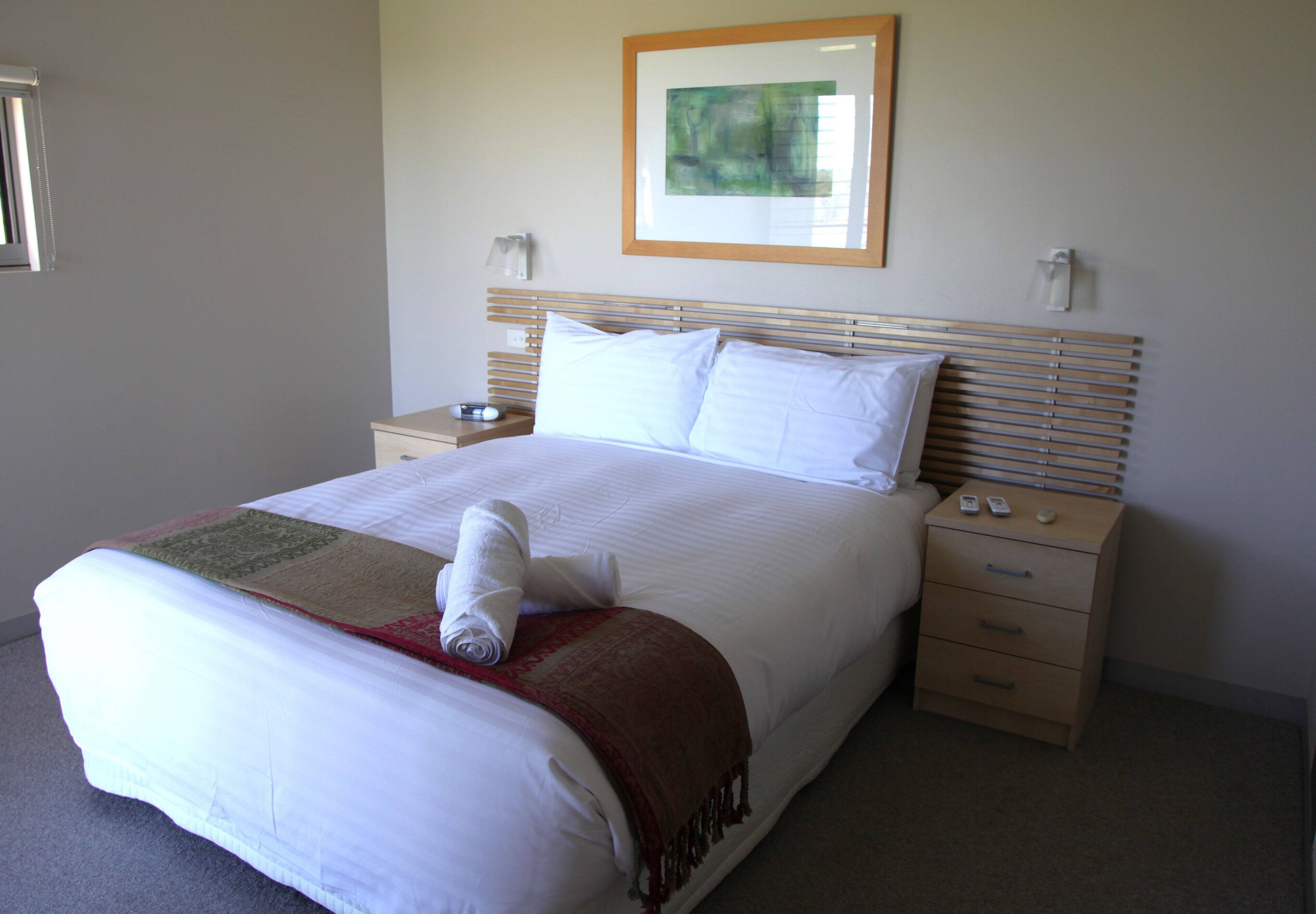 A neatly made bed with a white comforter and a red throw blanket, a wooden headboard, bedside tables with lamps, and a framed abstract painting on the wall.