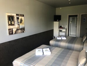 Ein Hotelzimmer mit zwei Betten, einem Fernseher auf einem Schrank, einem kleinen Schreibtisch mit Kaffeemaschine und einer Tür zu einem Badezimmer.