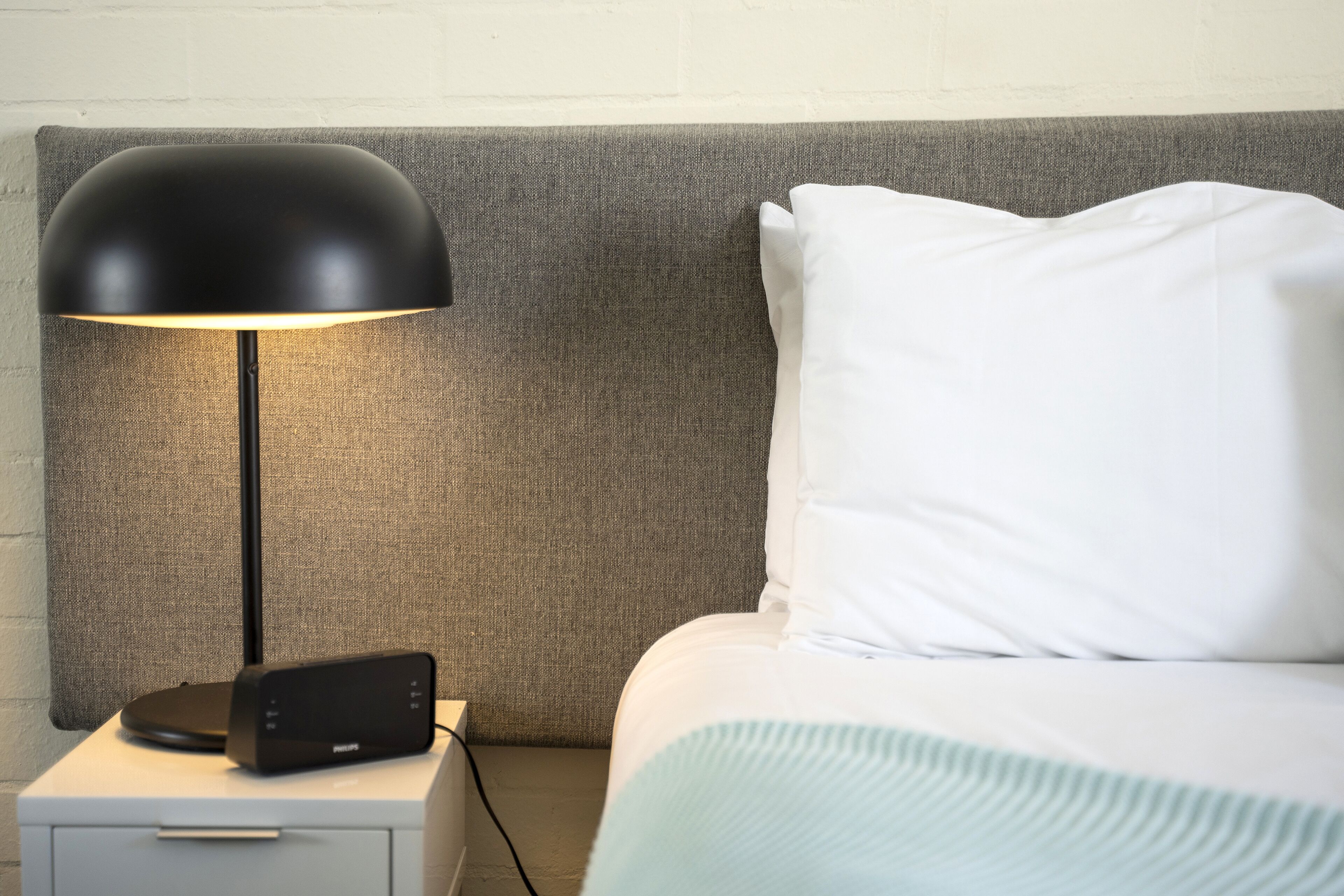 A neatly made bed with white pillows and a light blue blanket, a black lamp on a bedside table, and a Philips speaker.