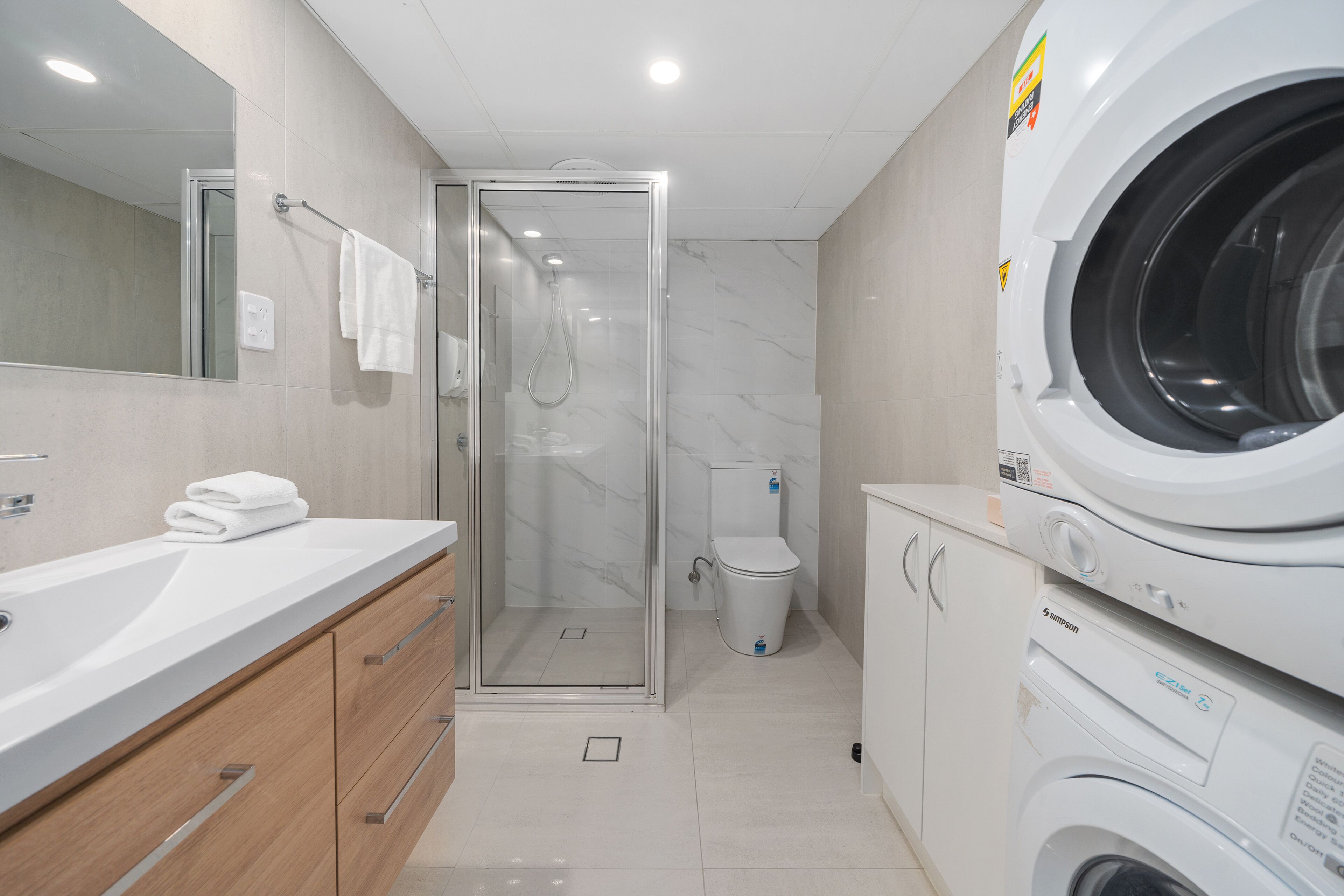 A modern bathroom with a glass-enclosed shower, a white sink with a mirror, and stacked washing machines.