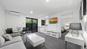 Una sala moderna con un sofá blanco, una mesa de centro, un mueble para TV y una lámpara. Hay un comedor con sillas y una mesa, y un dormitorio con una cama y una mesita de noche.