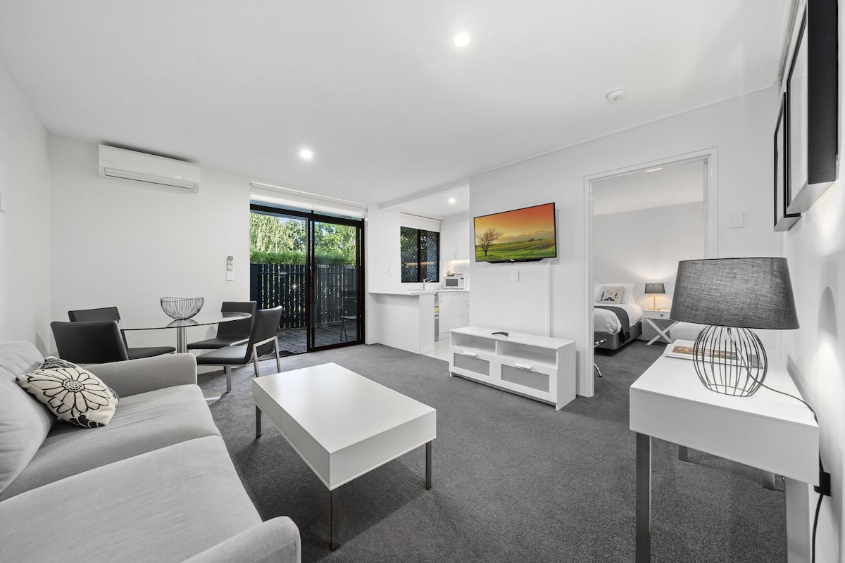 A modern living room with a white sofa, a coffee table, a TV stand, and a lamp. There is a dining area with chairs and a table, and a bedroom with a bed and nightstand.